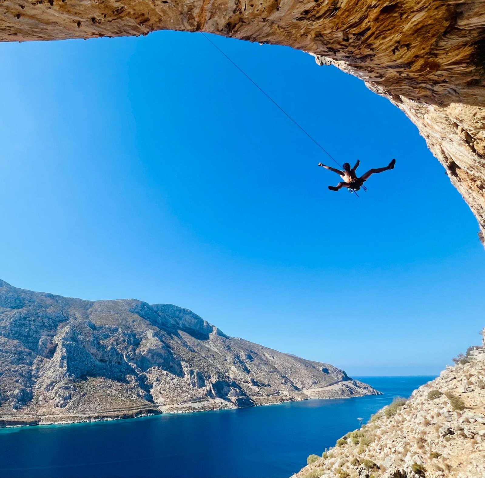 Une personne en chute libre dans un canyon rocheux au-dessus d'une mer ou d'un lac, sous un ciel clair et bleu.