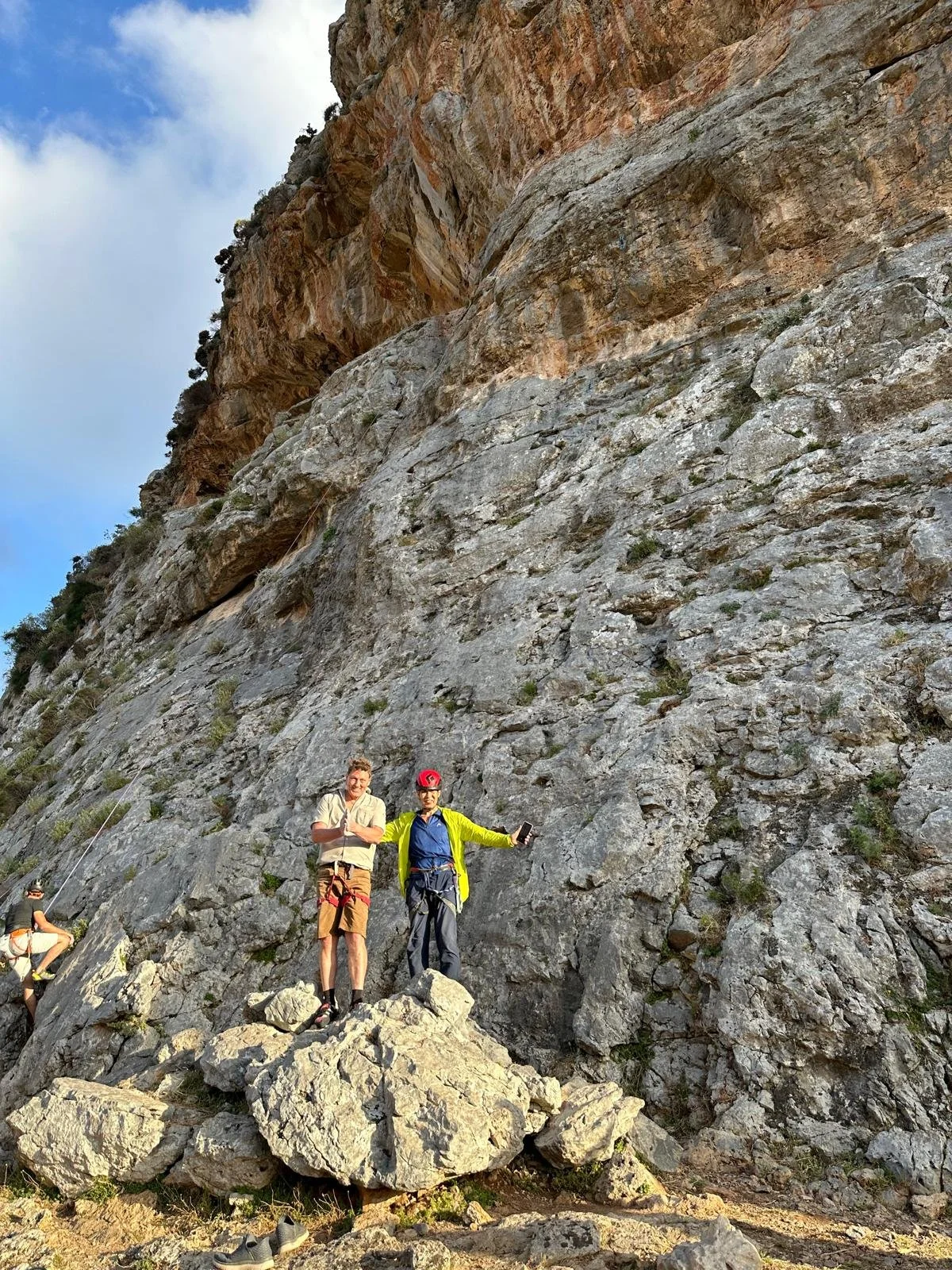 Deux hommes debout sur une pile de rochers devant une falaise rocheuse, un homme porte un casque rouge et un écran, l'autre porte une chemise beige avec ses bras croisés, et un troisième homme est partiellement visible à gauche, iconique d'une activi