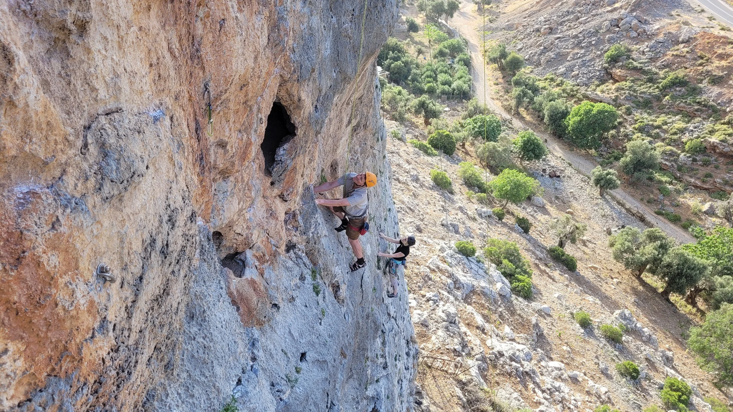 Deux grimpeurs escaladent une falaise rocheuse, avec un paysage désertique et quelques arbres en contrebas.
