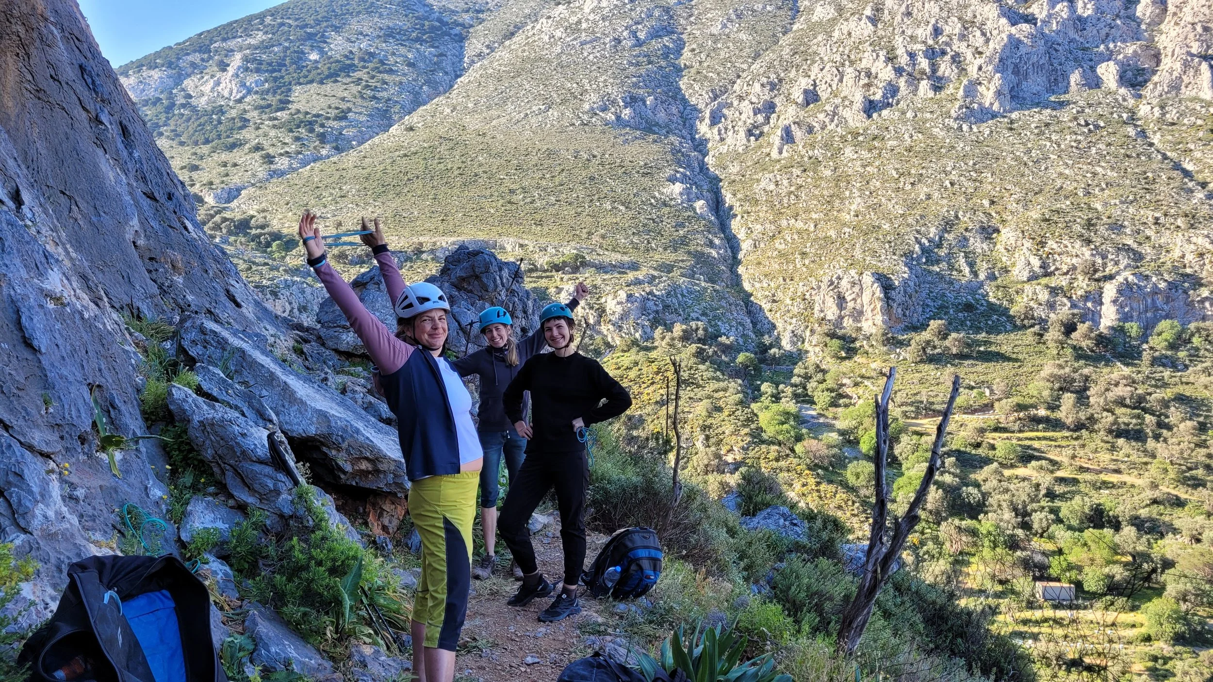 Trois femmes en randonnée en montagne, portant des casques, avec un paysage escarpé et verdoyant en arrière-plan.