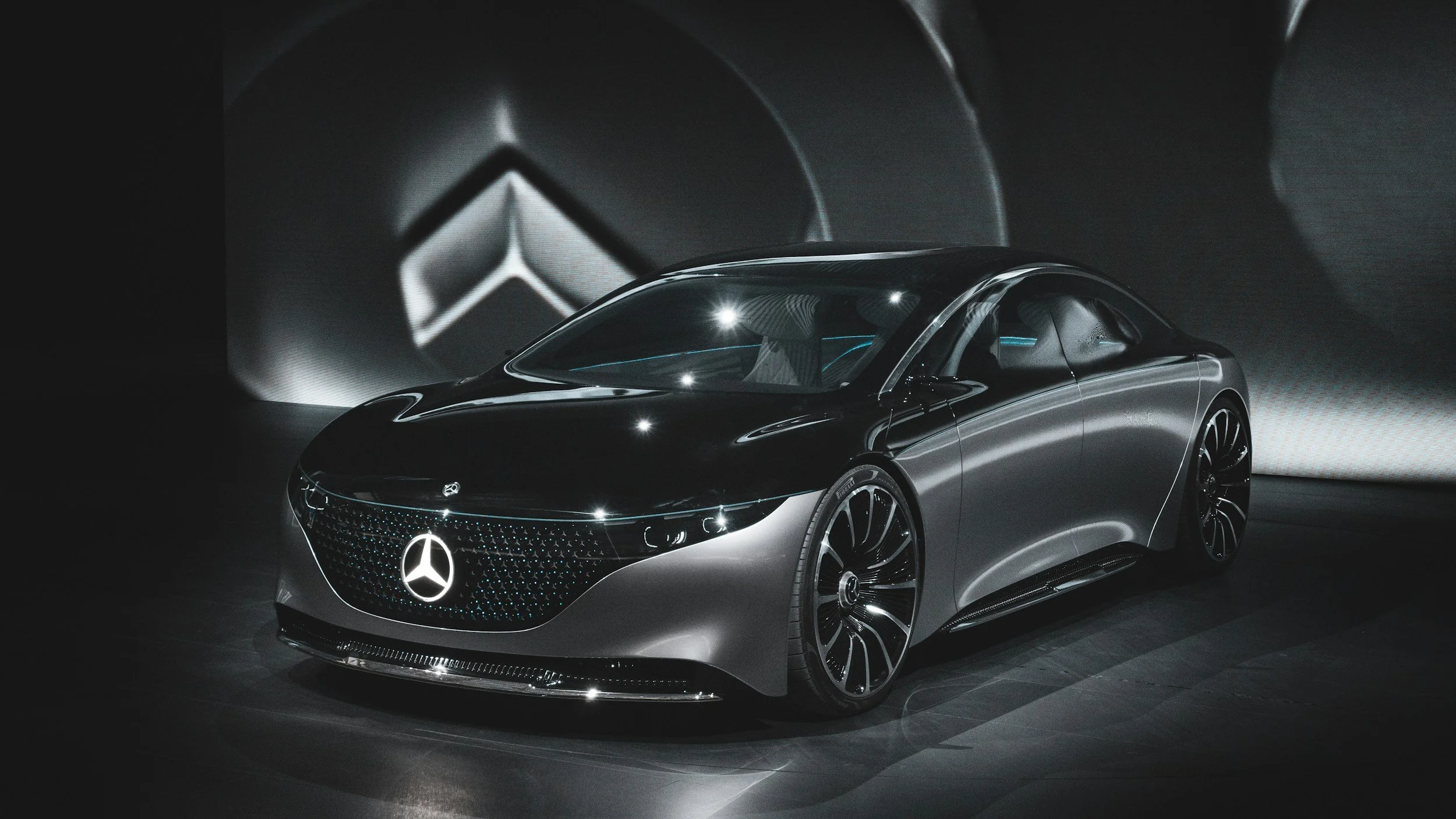 A sleek, futuristic Mercedes-Benz concept car with a glossy black and silver finish displayed at an auto show. The background features abstract geometric light patterns.