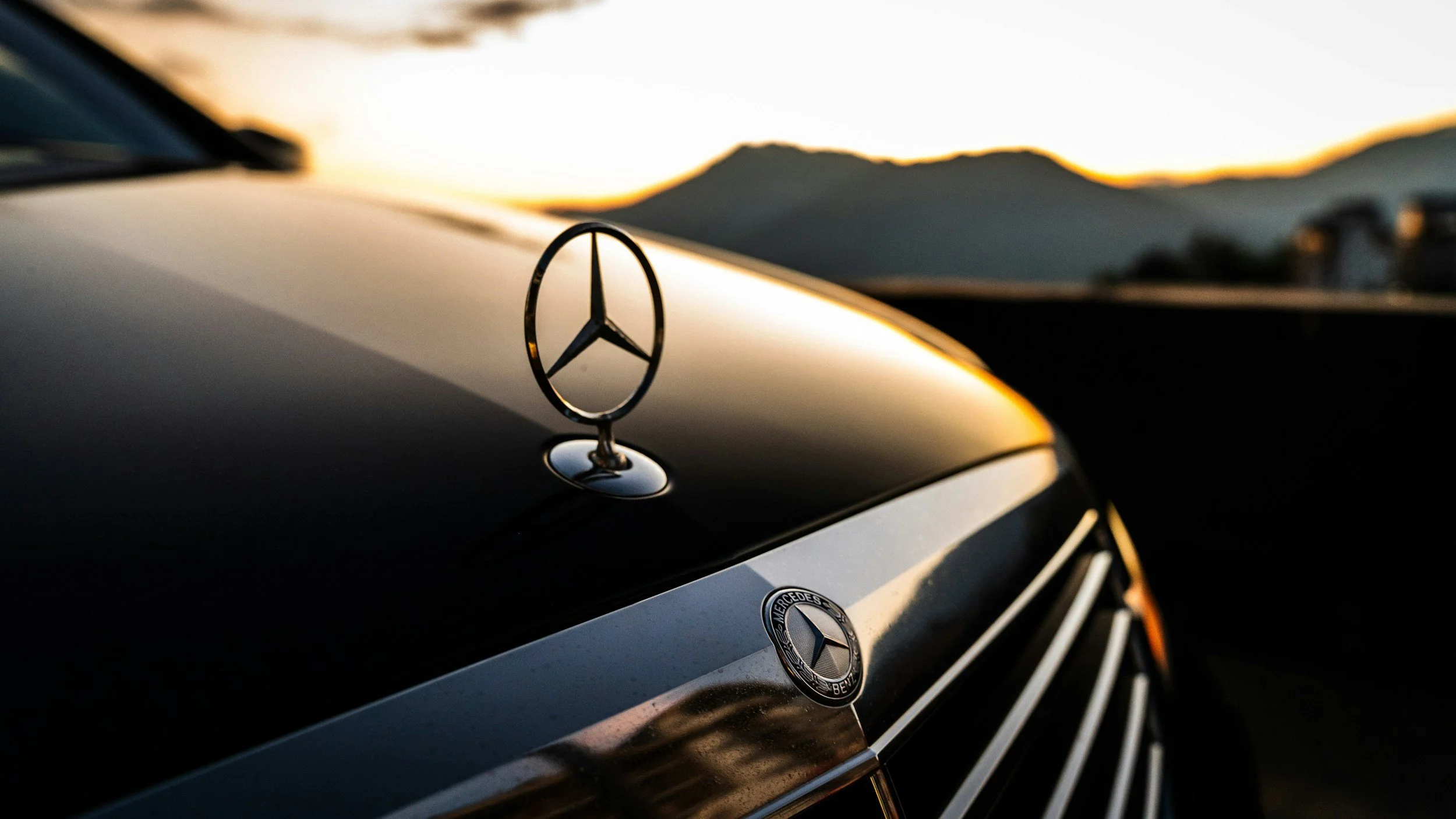 Close-up of a Mercedes-Benz emblem on the hood of a car during sunset with mountains in the background.