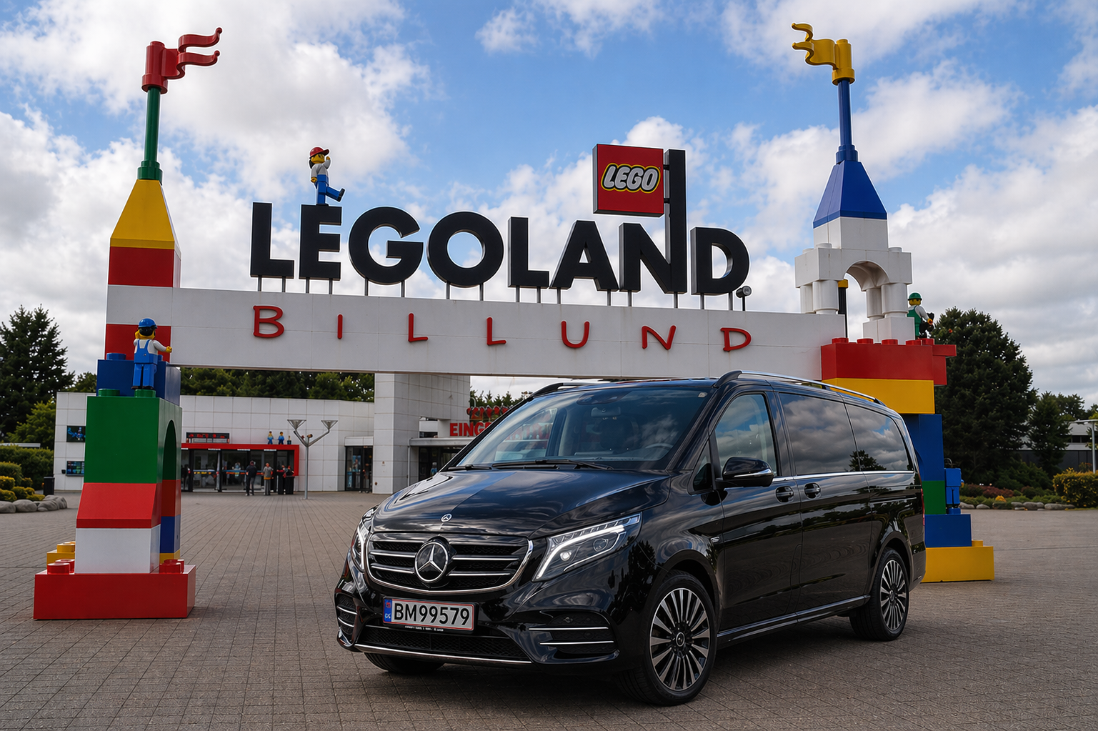 Entrance to Legoland Billund with large sign and Lego sculptures on top of pillars, a black Mercedes-Benz van parked in front, and a partly cloudy sky.