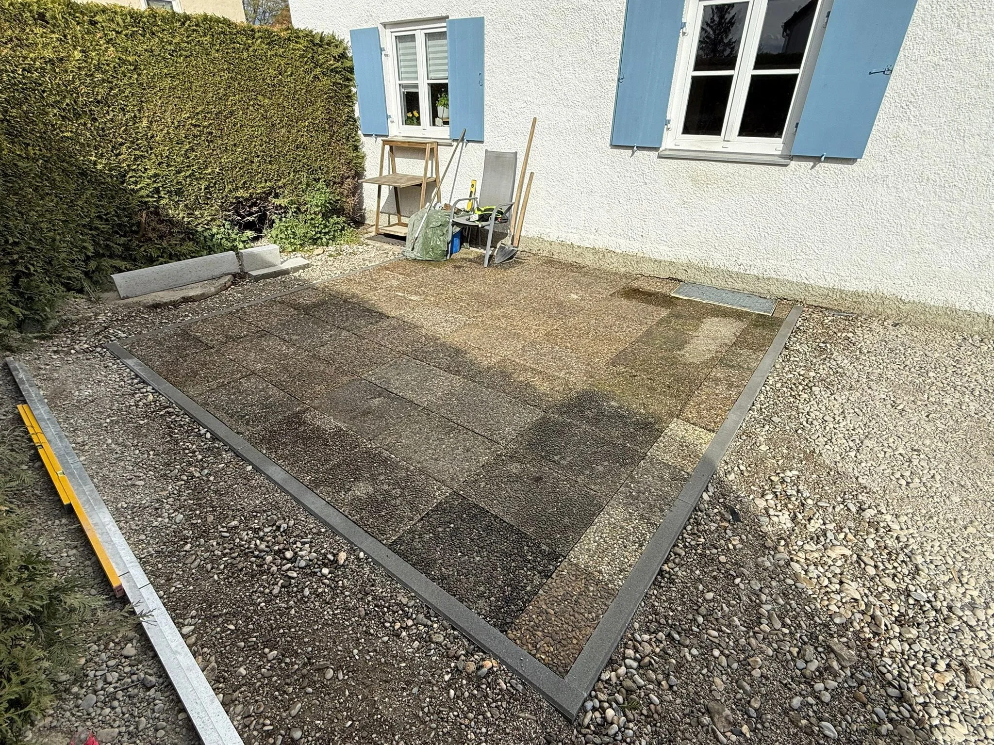 Ein geplanter Bereich für Pflasterarbeiten mit gepflasterten Steinen, umgeben von Kies und Gartenpflanzen, vor einem weißen Haus mit blauen Fensterläden.