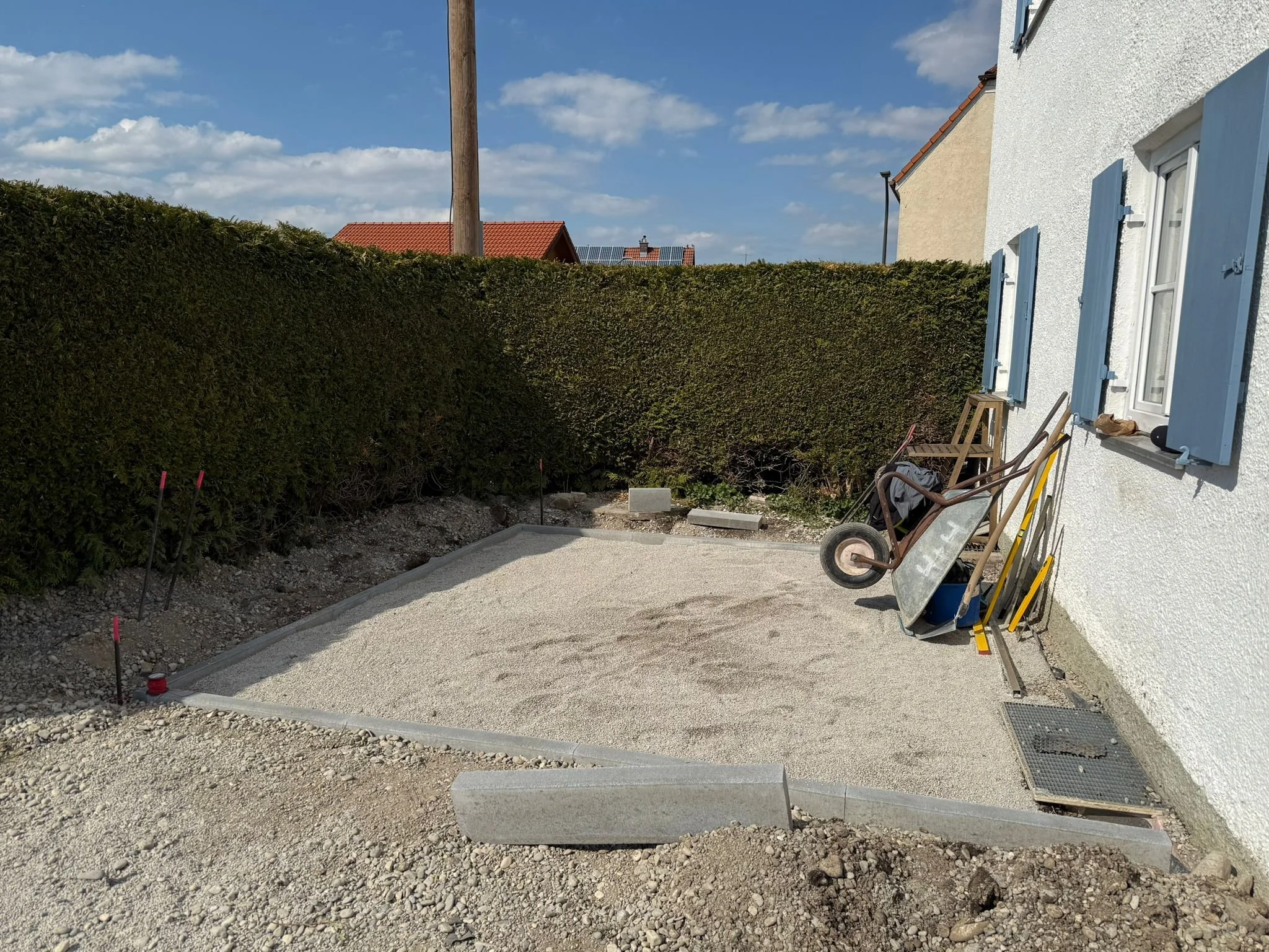 Gartenarbeit im Fortschritt: Ein Bereich neben einem weiß verputzten Haus mit blauen Fensterläden wird vorbereitet, mit Bauwerkzeugen, einer kleinen Schubkarre und Bastelarbeiten. Im Hintergrund ist ein hoher grüner Busch, und der Himmel ist blau mit