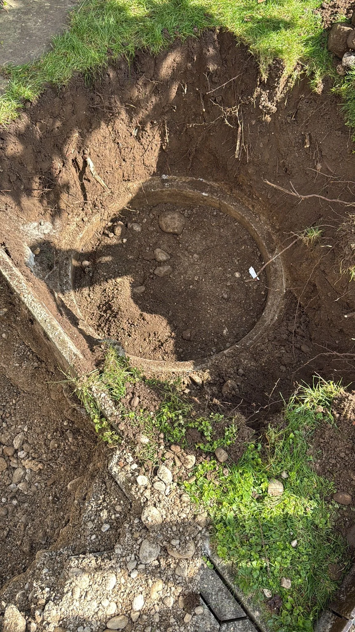 Ein einsturzgefährliches Erdloch wurde ausgehoben und mit einer runden, alten Betonröhre oder einem Meterrohr versehen, vermutlich für Installationen oder Brunnen. Blätter und Gras umgeben die Grube, und die Umgebung scheint eine Baumaßnahme oder War