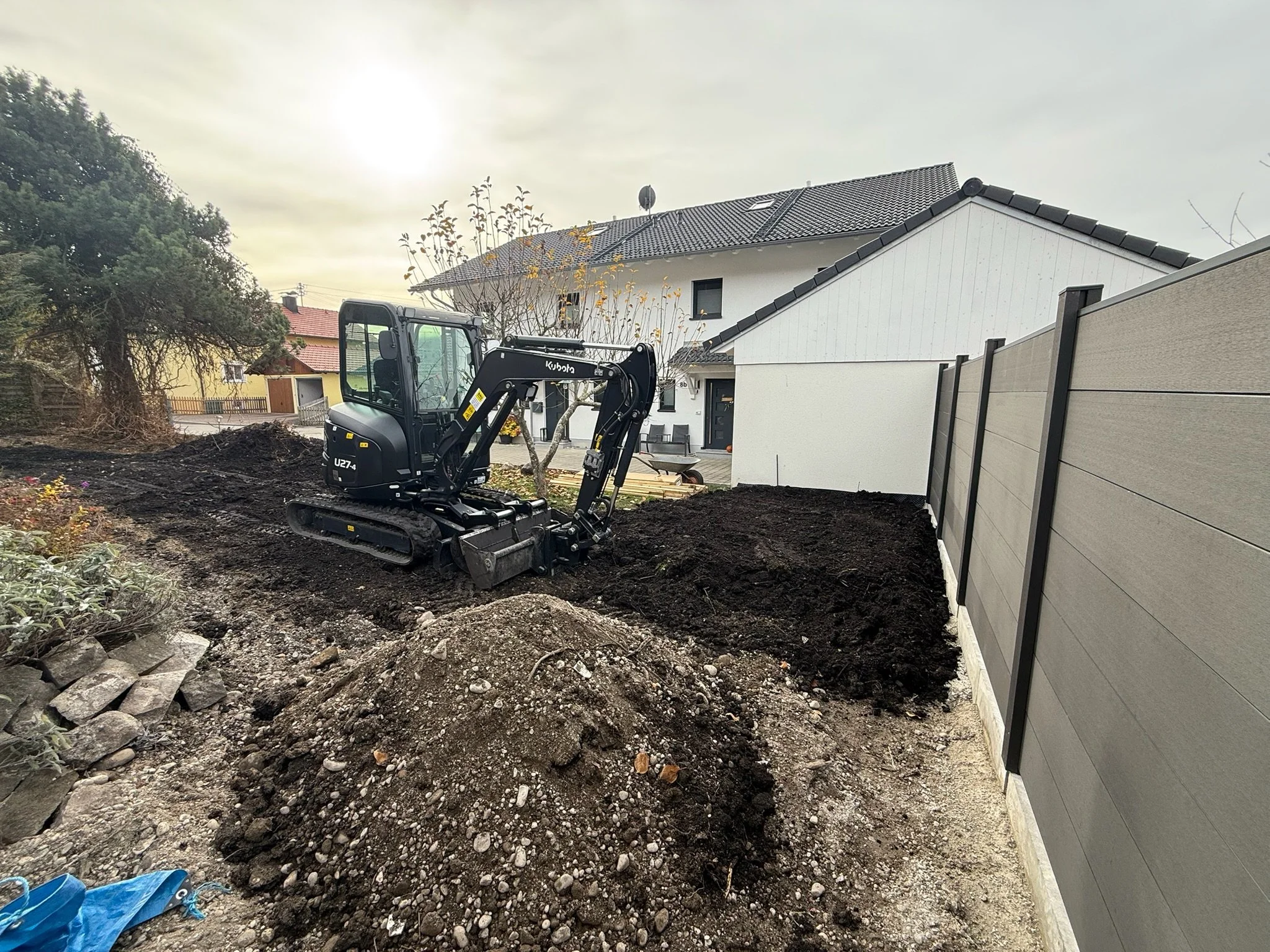 Ein kleiner Bagger arbeitet in einem Garten neben einem Hochzaun bei Tageslicht.