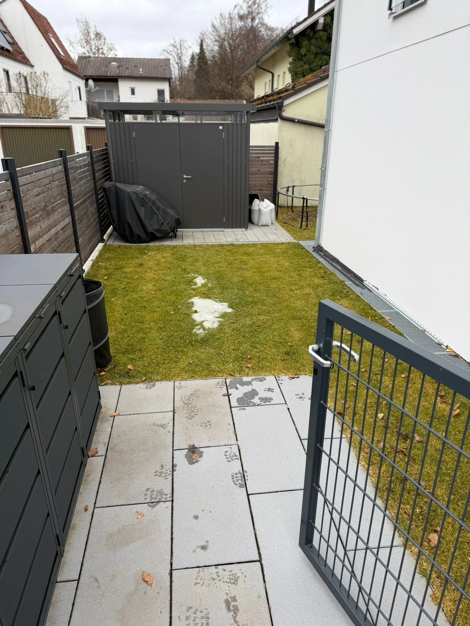 Blick auf einen kleinen Garten mit gepflastertem Weg, Gras und einem kleinen Schuppen. Es sind Fußspuren im nassen Sand zu sehen.