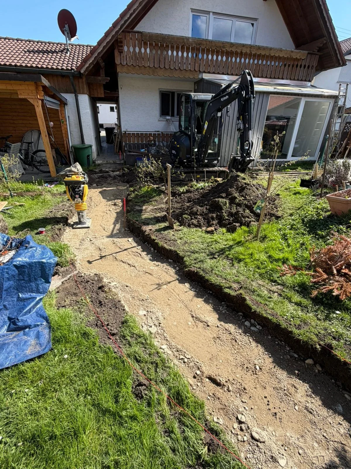 Garten wird mit einer kleinen Baggermaschine umgestaltet, ein Weg wird vorbereitet. Es sind Werkzeuge und Baumaterialien sichtbar, während im Hintergrund ein Haus steht.