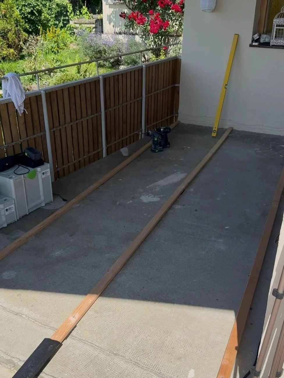 Ein Balkon im Bau oder in Renovierung, mit Holzlatten, einem Akkuschrauber, einer Wasserwaage, Müll und Werkzeug, sowie einem Sichtschutz aus Holz und Gartenpflanzen im Hintergrund.