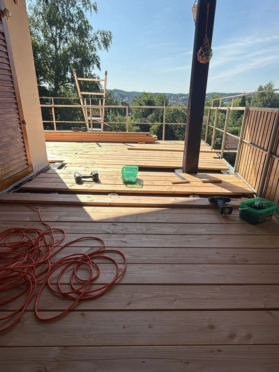 Der Balkon ist im Bau und es liegen Bauwerkzeuge und Holzbohlen auf dem Boden. Im Hintergrund sind Bäume und ein blauer Himmel zu sehen.