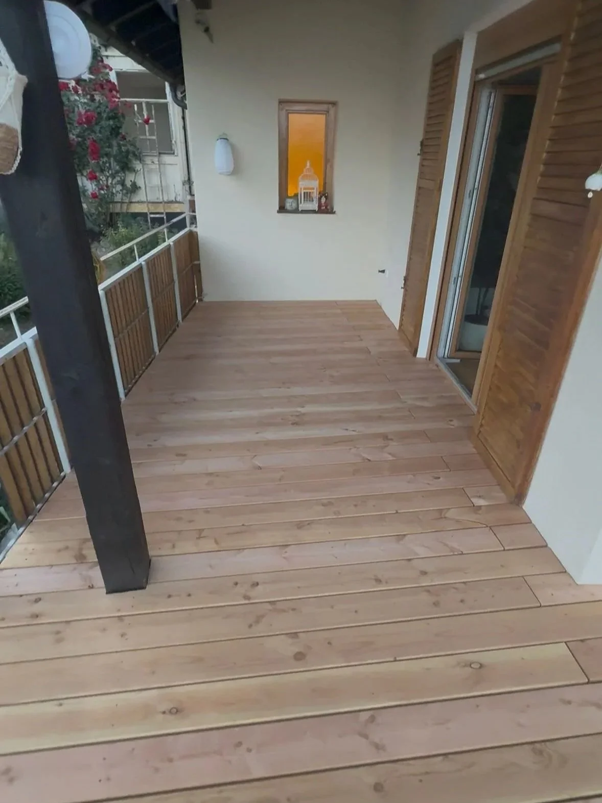 Blick auf eine Terrasse mit Holzdielen, eine weiße Wand mit einem kleinen Fenster, eine schiebbare Tür, Laterne und Dekorationen im Fenster, und eine Haltese für Pflanzen oder Gegenstände auf der linken Seite.