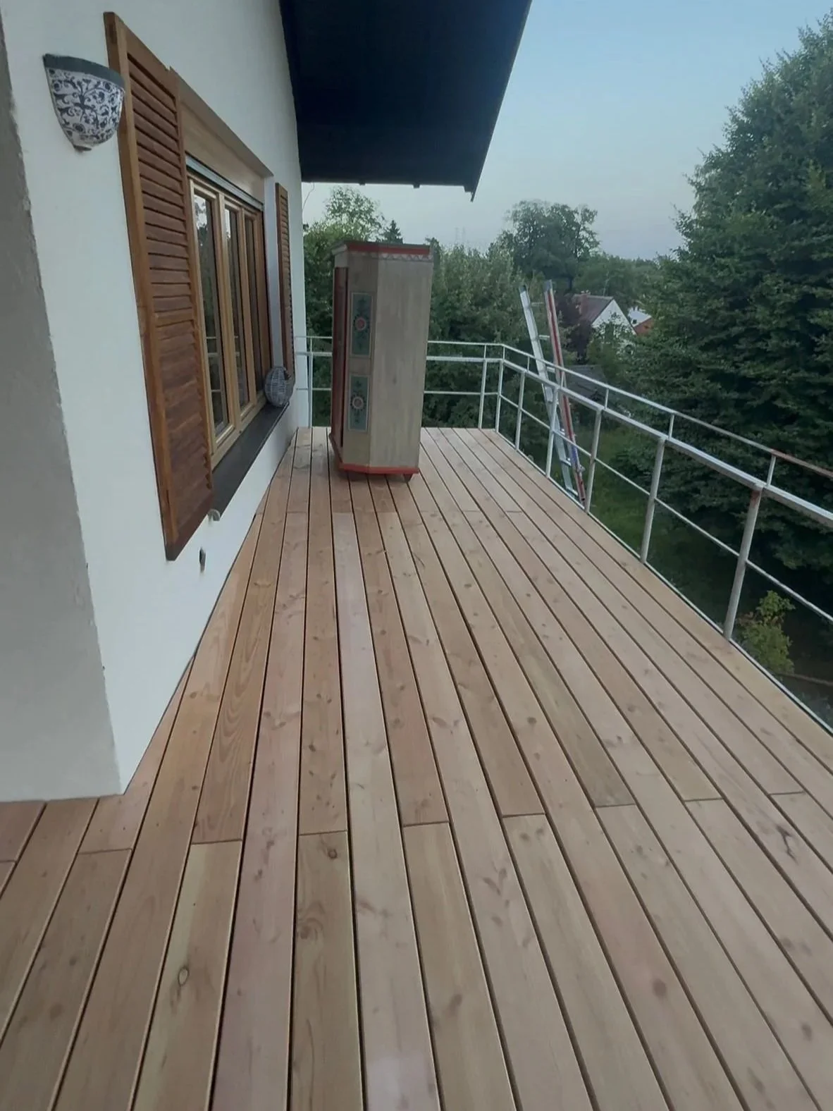 Blick auf eine große Balkon mit frisch verlegtem Holzboden, Fenster mit Holzläden und Metallgeländer, umgeben von Bäumen und Häusern in der Ferne.
