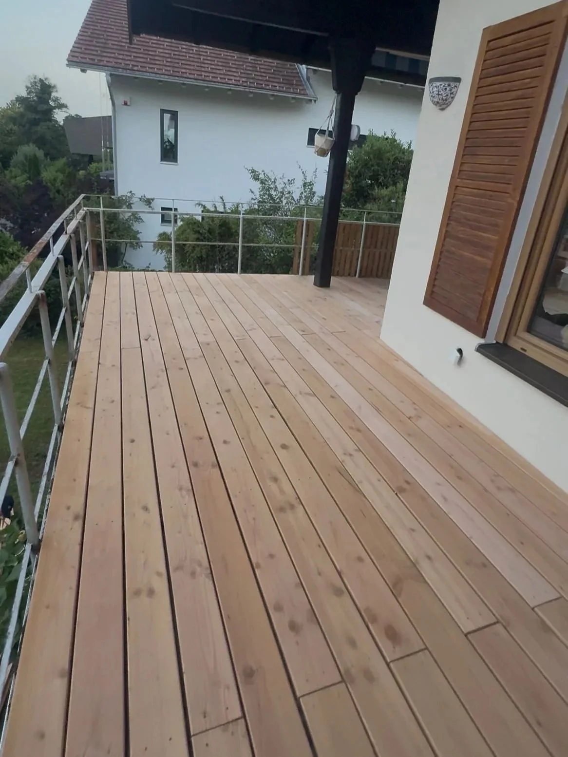 Neue Holzterrasse vor einem Wohnhaus, mit geschlossenem Fenster und Garten im Hintergrund.