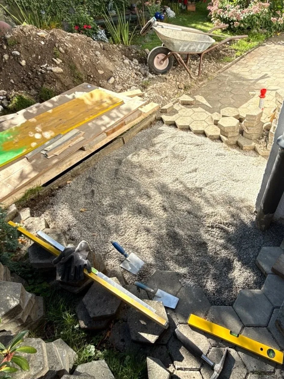 Gartenweg wird mit Pflastersteinen gebaut, Schaufel, Wasserwaage und Handschuhe sind sichtbar, Baustelle mit Kies, Steinen und einer Schubkarre im Garten.