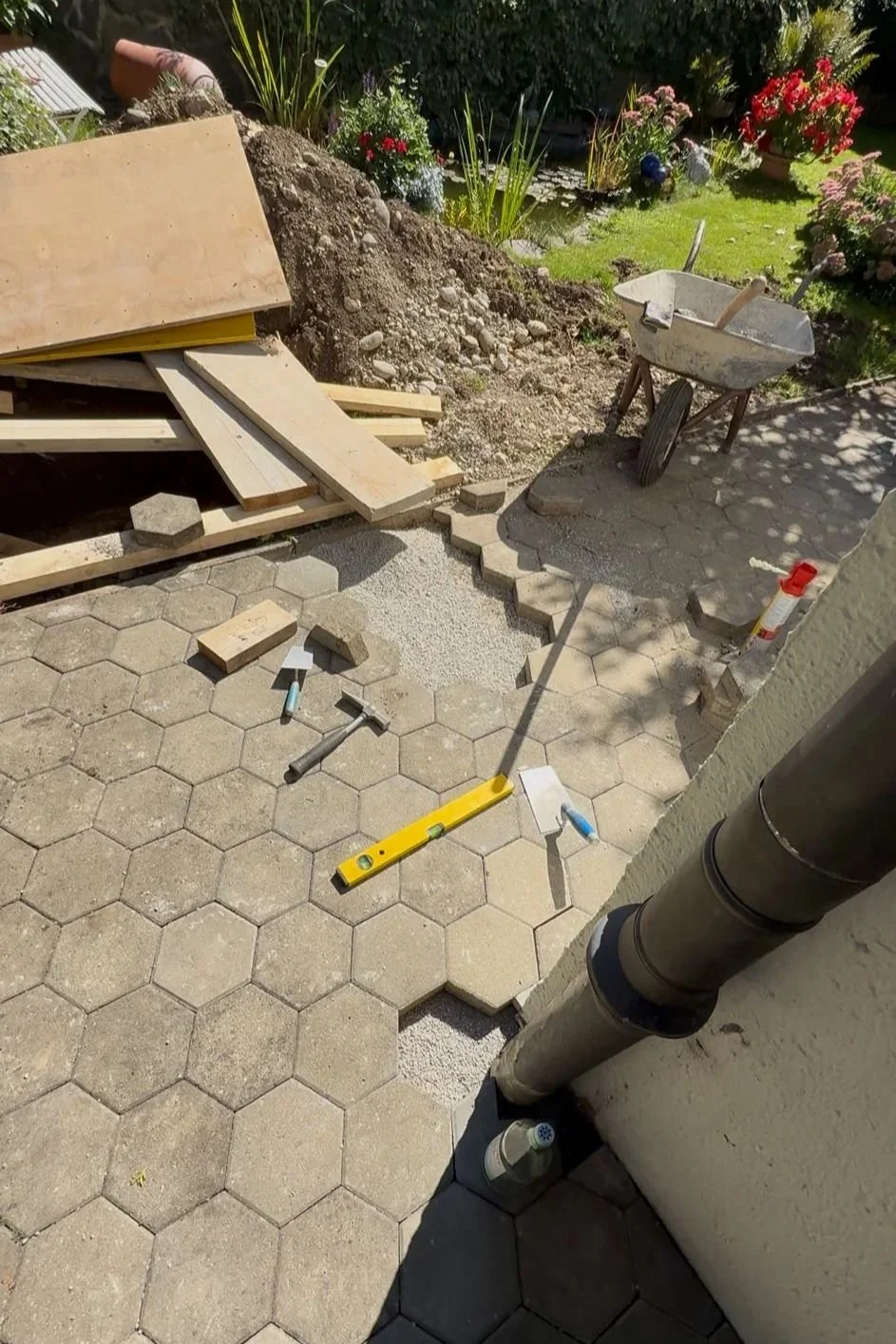 Der Garten wird mit Pflastersteinen befestigt, wobei Werkzeuge wie Wasserwaage, Hammer und Fugenkreuze sichtbar sind. Es gibt einen Schubkarren, Sandhaufen, Holzbohlen und eine Blumenterrasse im Hintergrund.