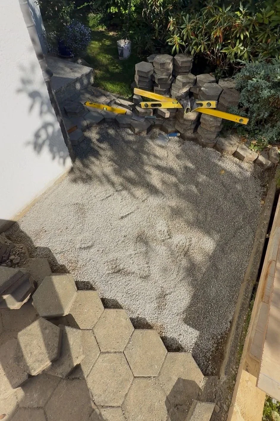 Ein Hausgarten mit Fundamentarbeiten. Es liegen Pflastersteine, ein Wasserwaage und Steine zum Bau einer Beeteinfassung. Es ist Gartenlaubenerde, und ein Schatten einer Pflanze ist sichtbar.