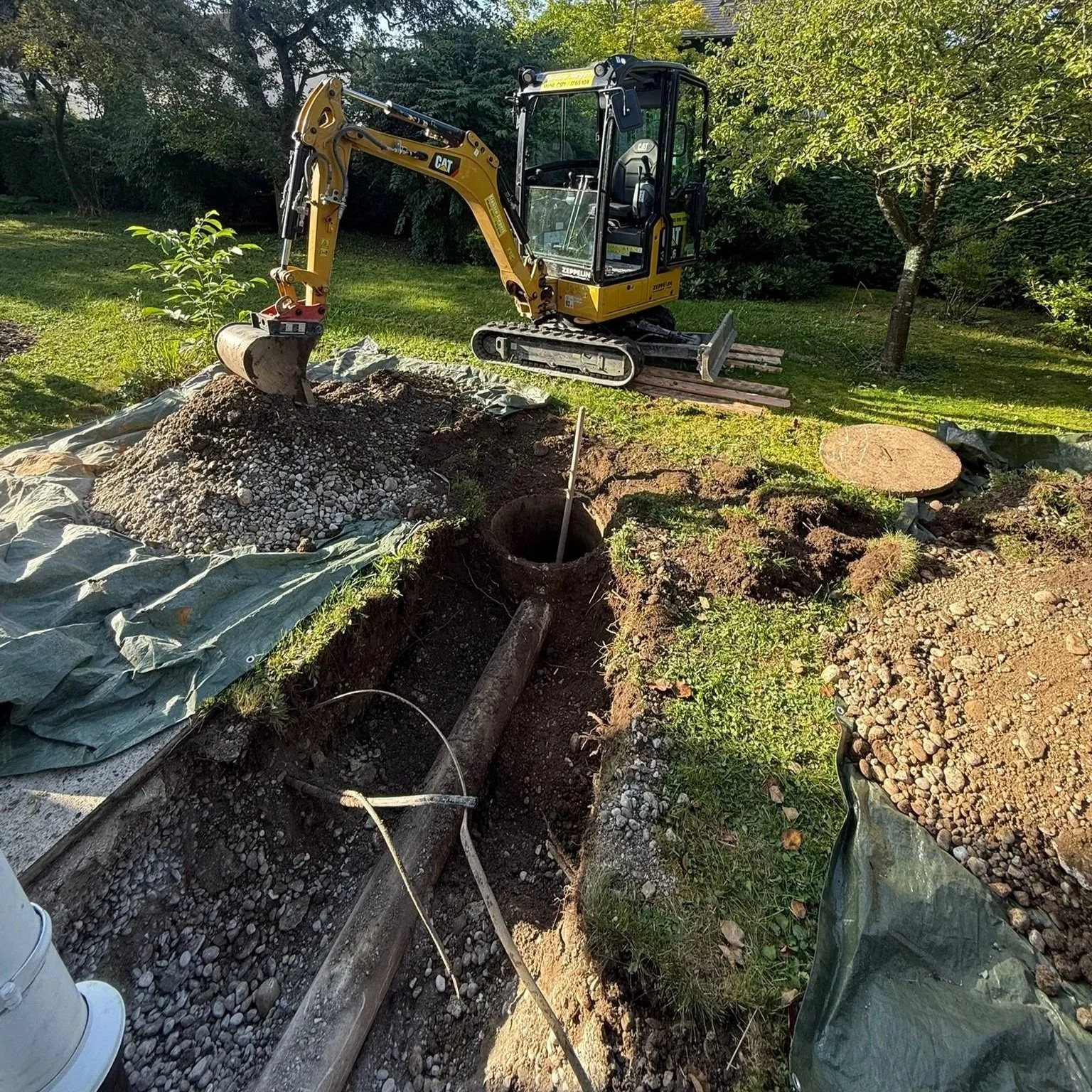 Mini-Bagger arbeitet an einer Wasserleitung in einem Garten mit Bäumen und Gras.