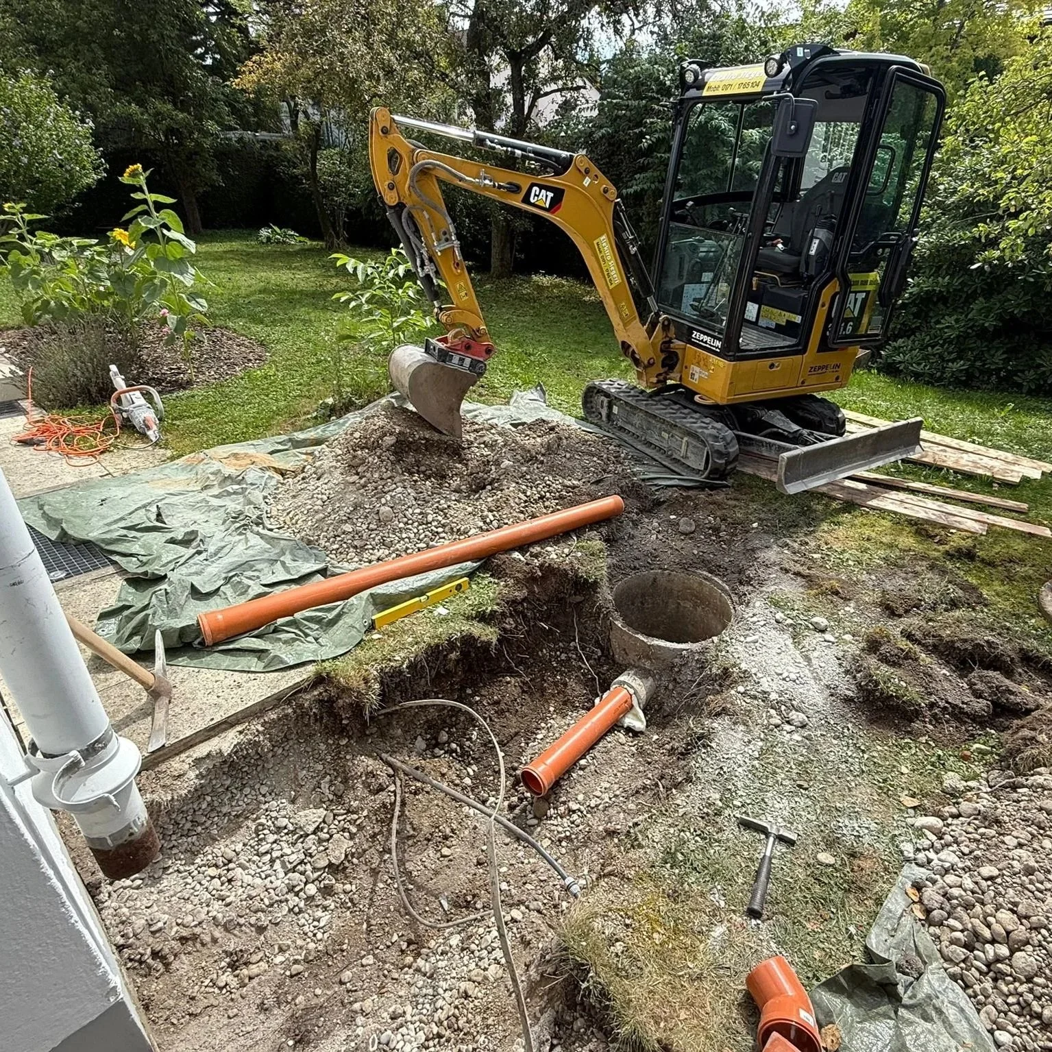 Mini-Bagger beim Aushub eines Grabens für Rohrleitungen in einem Garten mit Bäumen und Sträuchern im Hintergrund.