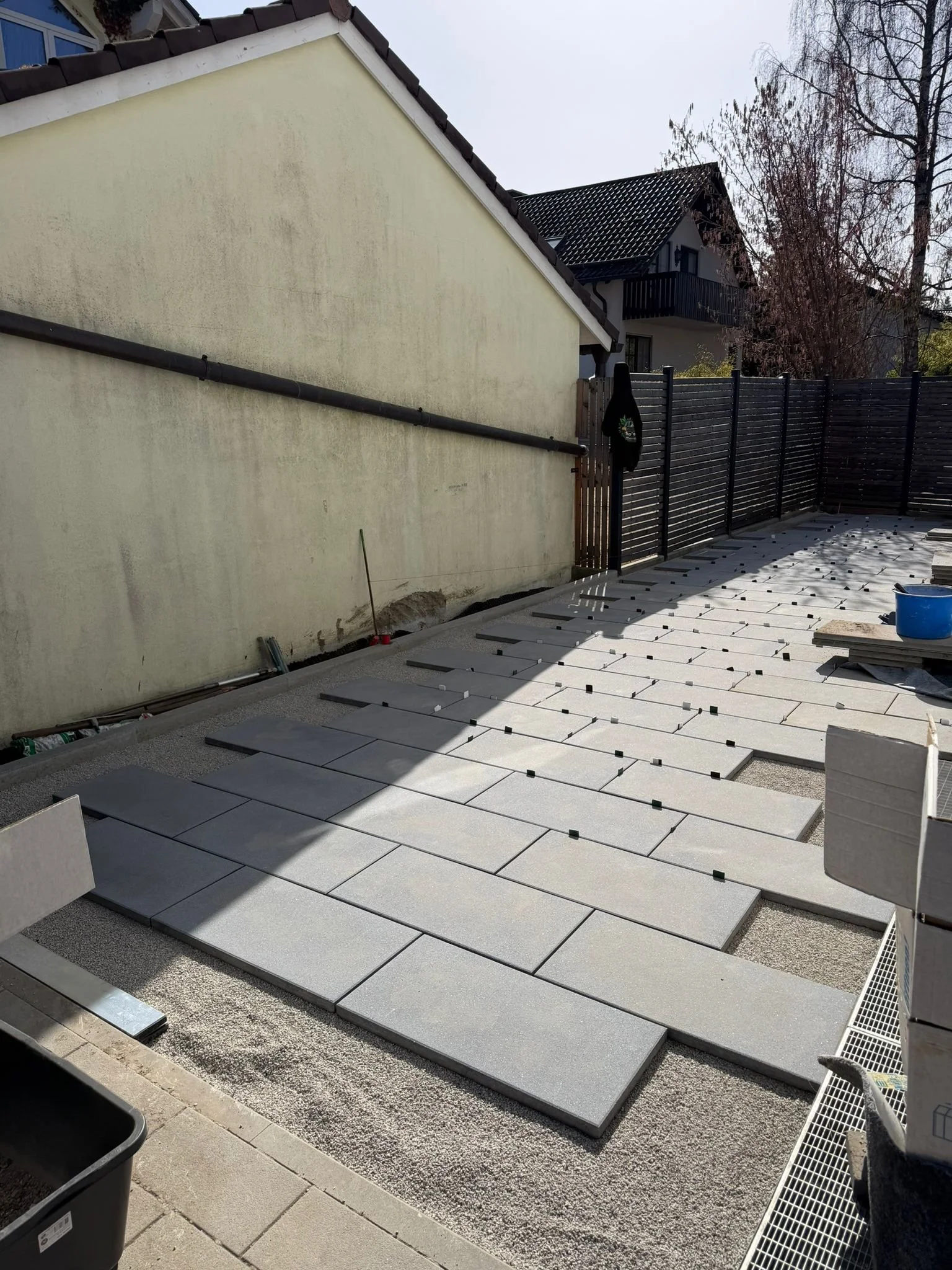 Ein Garten mit frisch verlegten Plattensteinen auf der Terrasse, im Hintergrund ein Zaun, eine Hauswand und ein zweistöckiges Haus mit Balkon. Es scheint, dass die Terrasse renoviert oder neu gestaltet wird.