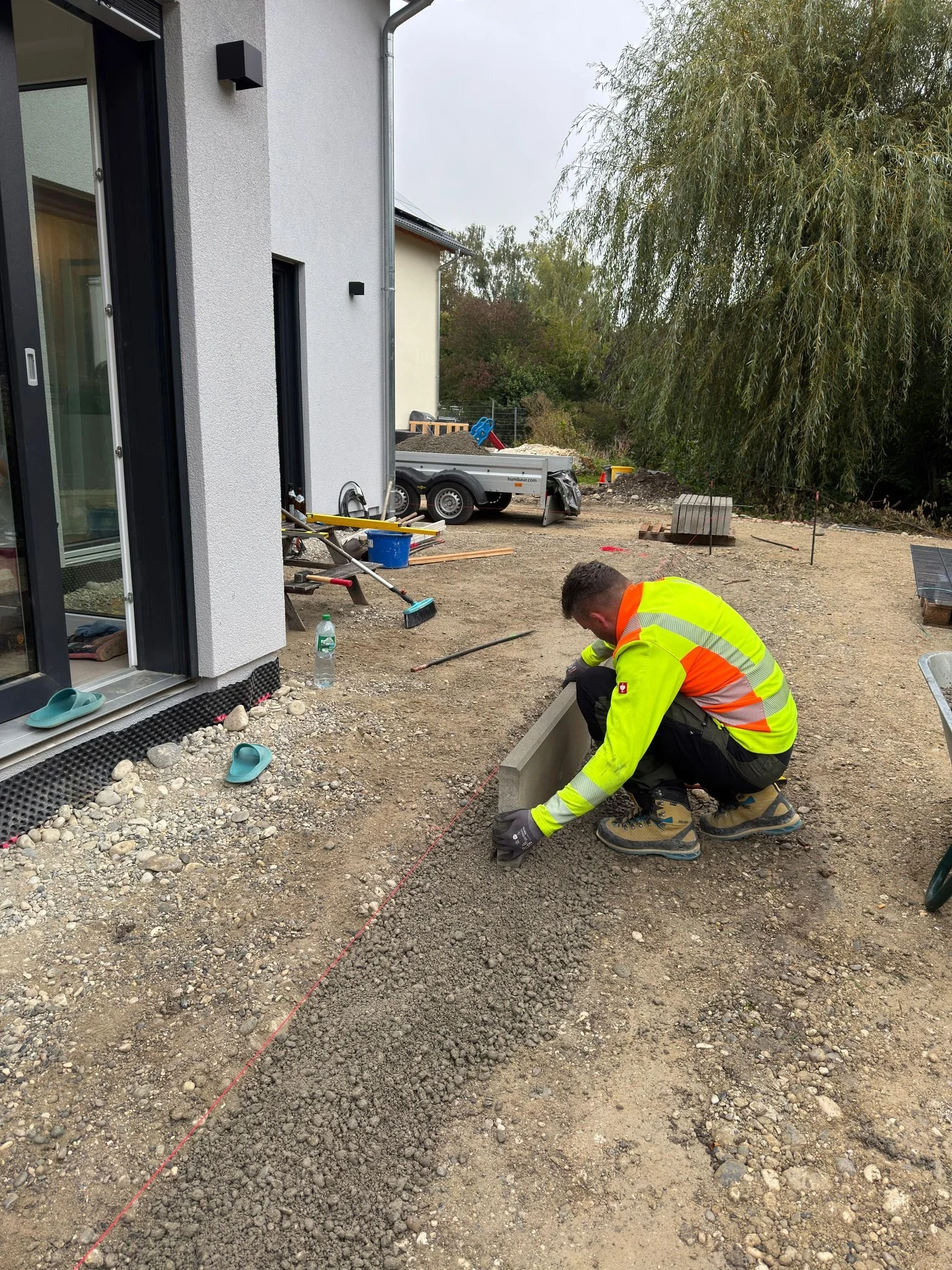 Ein Bauarbeiter arbeitet auf einer Baustelle vor einem modernen Haus, er legt eine Reihe von Pflastersteinen im Gartenbereich.