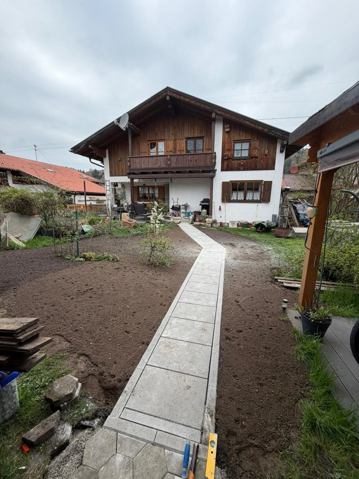 Garten mit neu gepflastertem Weg zur Hausesitzung im ländlichen Stil, mit Gartenmöbeln, Blumentöpfen und Gemüsebeeten, im Hintergrund ein Holzhaus mit Balkon.