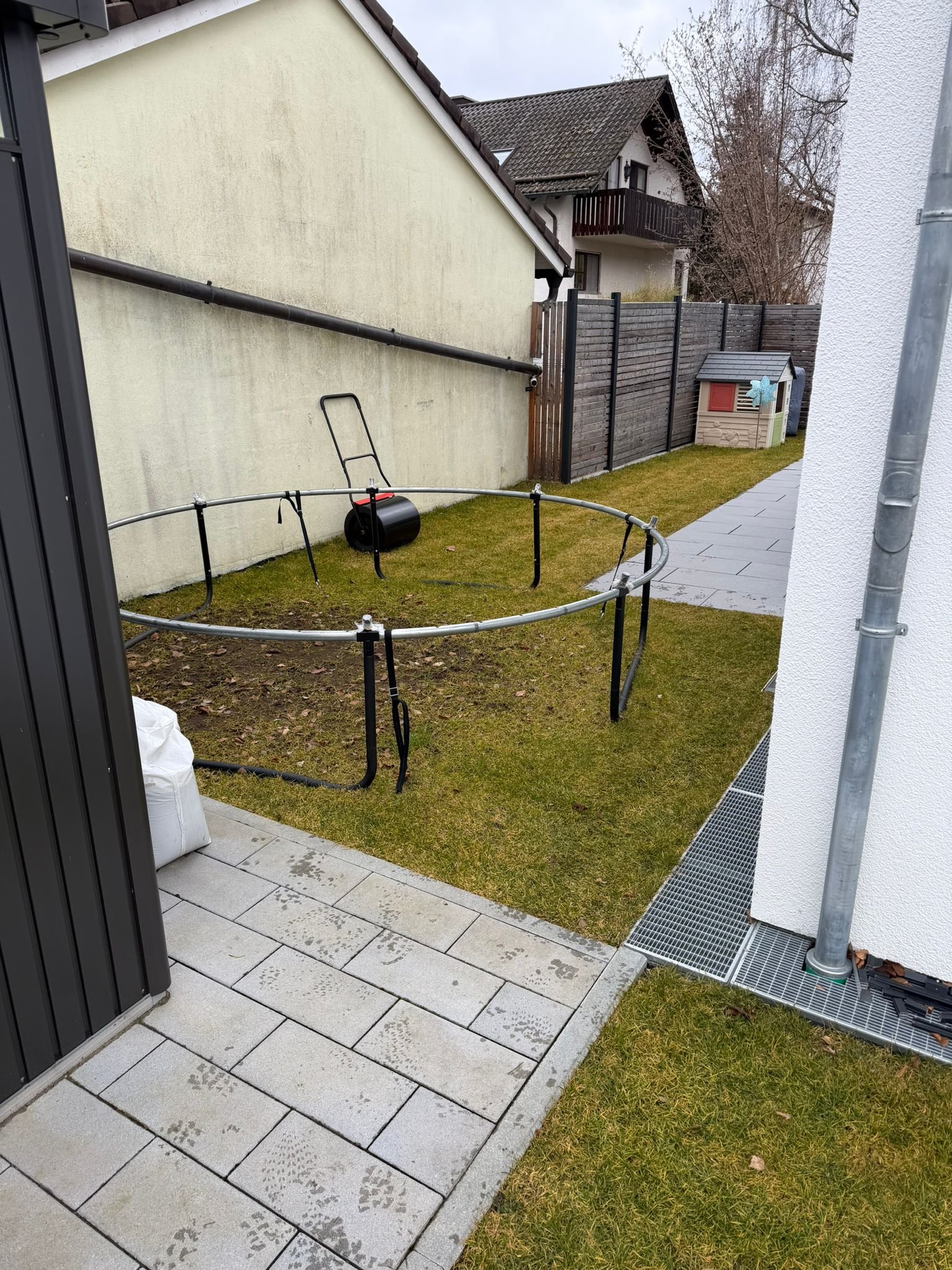 Garten mit Gras, gepflastertem Weg, Gartenhäuschen, leere Trampolin, Zaun und Haus im Hintergrund