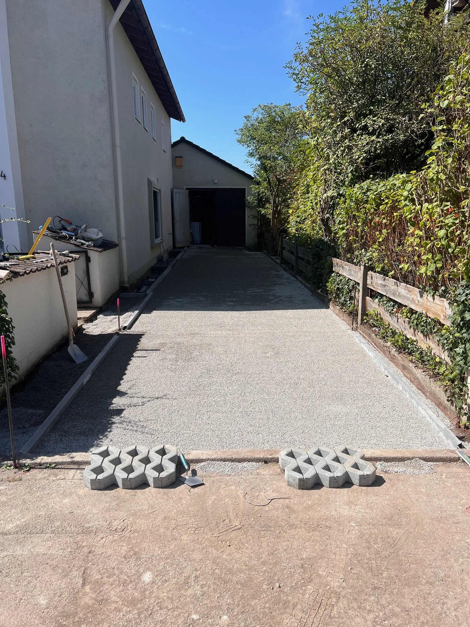 Ein frisch gepflasterter Gehweg vor einem Haus, umgeben von Bäumen und Zäunen auf der rechten Seite, mit Baumarktmaterialien im Vordergrund.