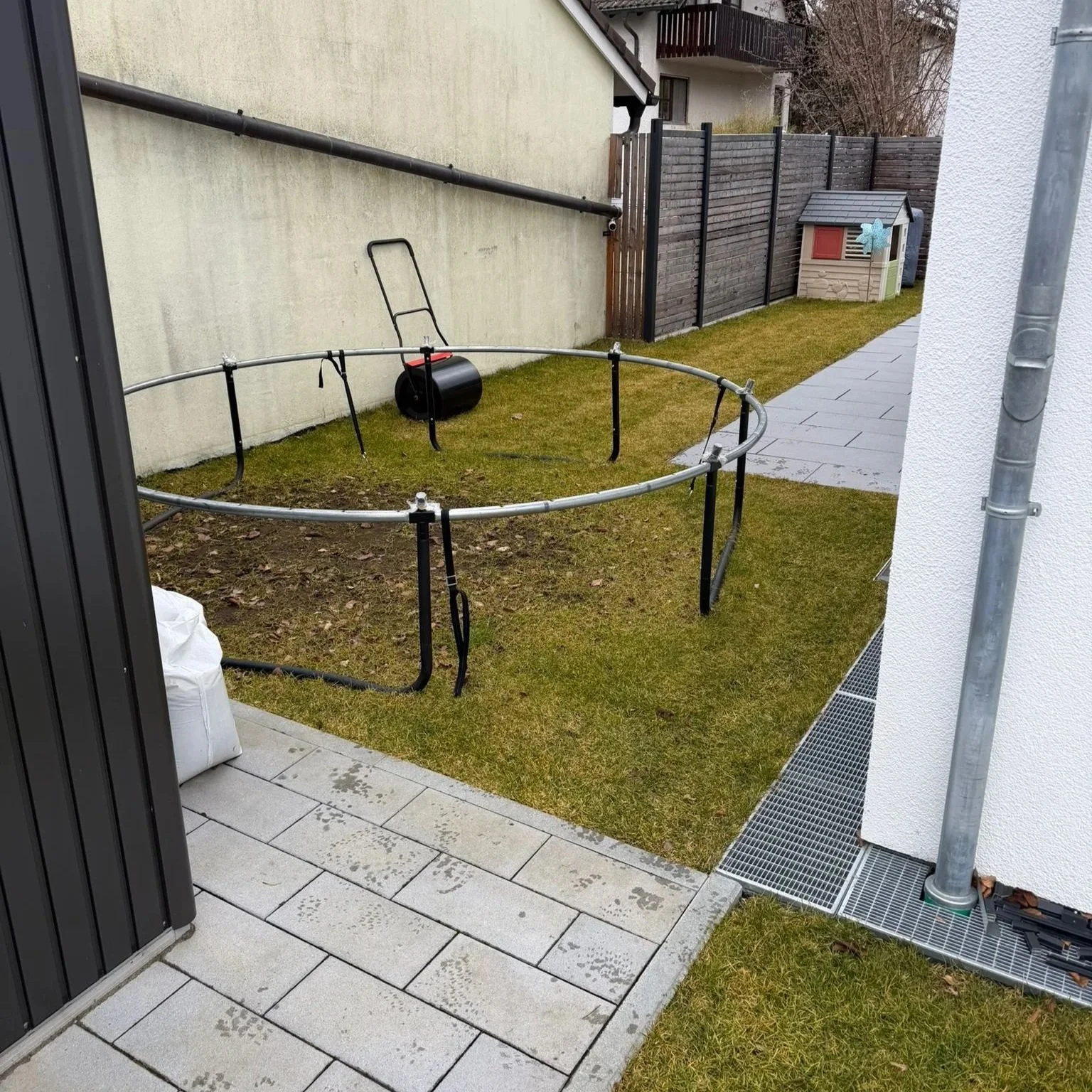 Ein Garten mit Rasen und einem umgedrehten Trampolin, ein Rasenmäher und ein Gartenhäuschen im Hintergrund. Es gibt einen Kiesweg und einen Zaun.