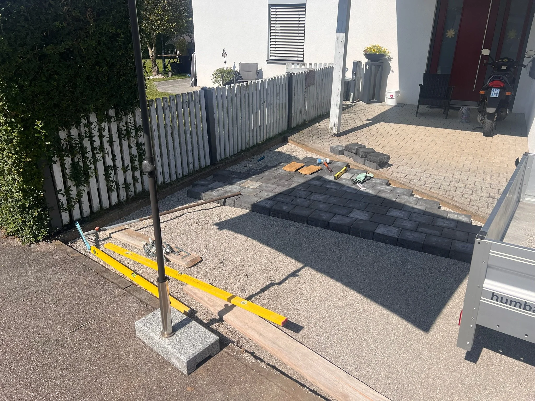 Der Gehweg wird gerade gepflastert, mit Baustellenausrüstung und Steinen, im Hintergrund eine Hausfassade mit Garten und Stellplatz.