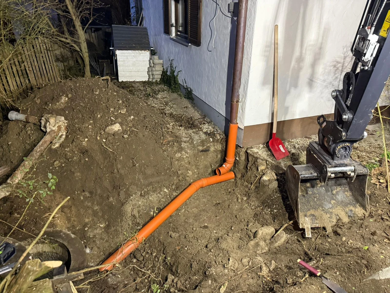 A construction site neben einem Haus mit einem Erdaushub, einem orangefarbenen Abwasserschacht, einer Schaufel und einem Baugerät.