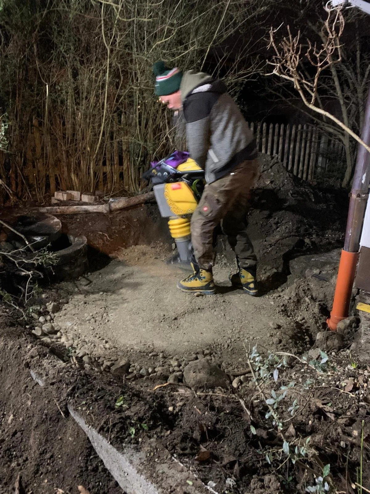 Ein Mann arbeitet nachts an einem Garten- oder Landschaftsbauprojekt, während er eine Maschine oder Werkzeuge benutzt, um den Boden vorzubereiten.