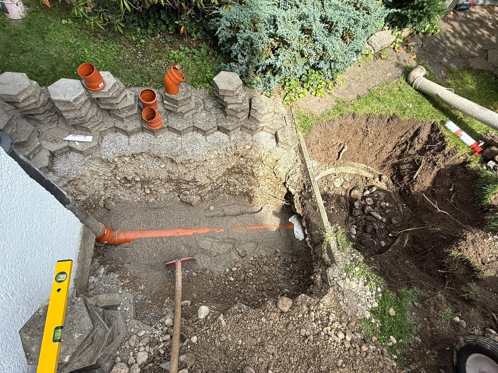 Graben für Wasserleitung oder Rohrleitung im Garten, mit Rohren und Werkzeugen. Bunte Rohre auf dem Boden und im Graben, Steine und Erde um den Graben herum, im Hintergrund Pflanzen und Sträucher.