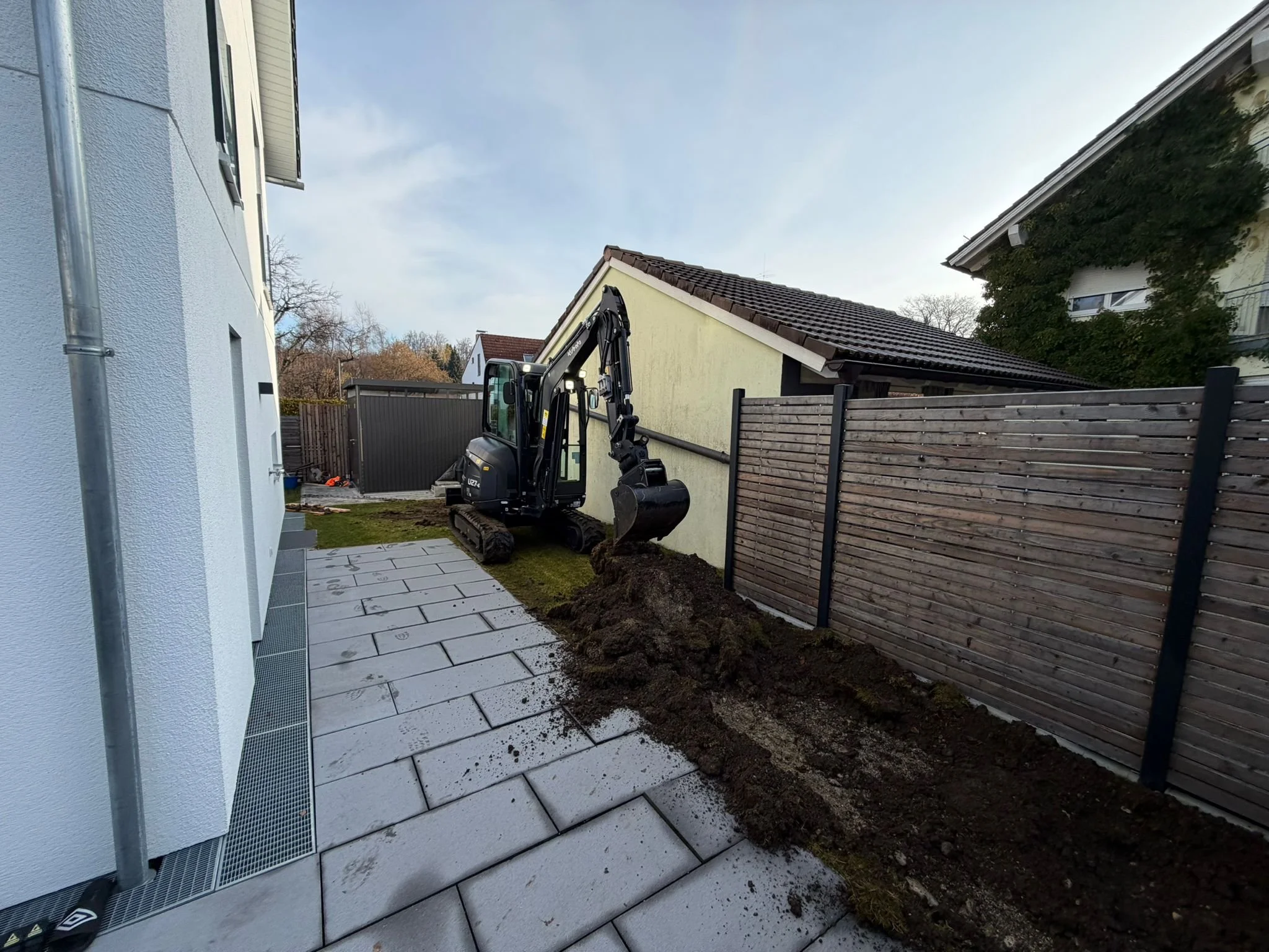 Baustelle im Garten mit einem kleinen Bagger, der Erde bewegt, neben neu verlegten Pflastersteinen und einem Zaun.