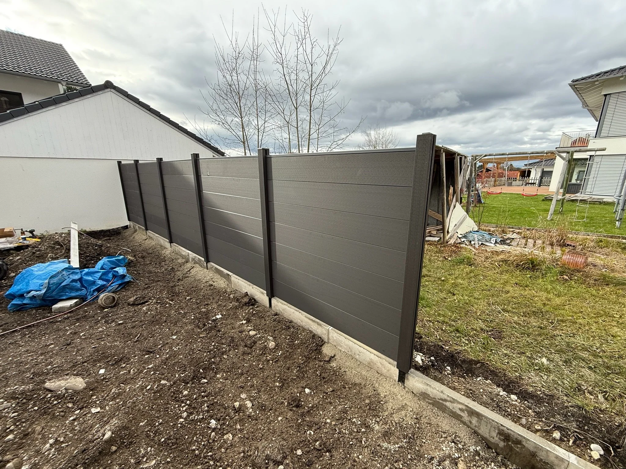 Neuer grauer Sichtschutzzaun aus Kunststoff oder Holz in einem Garten, mit Bauarbeiten und Werkzeug im Vordergrund, auf der linken Seite ein Teil des Hauses, am Himmel dunkle Wolken.