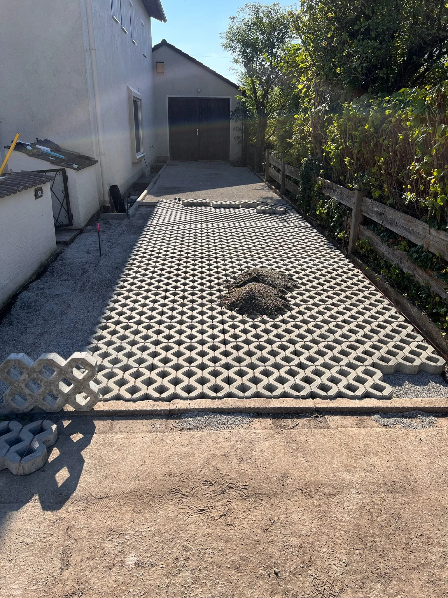 Der Vorgartenbereich mit Pflastersteinen, einige lose Pflastersteine und Trödel im Bereich links. Der Übergang zur Einfahrt ist sichtbar, im Hintergrund ein Gartentor und Bäume. Auf der rechten Seite eine Holzzäunung mit Kletterpflanzen.
