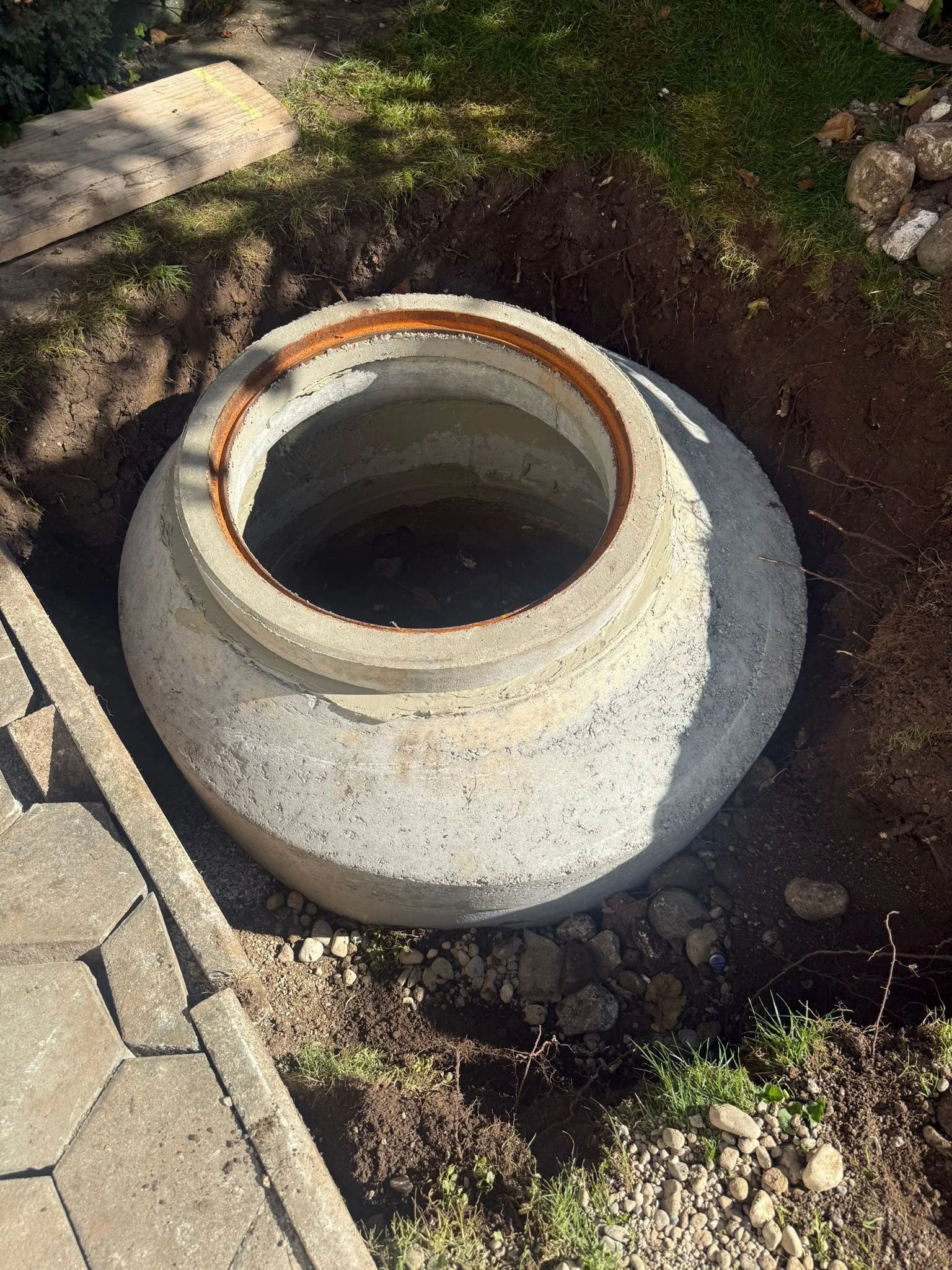 Große runde Betonzisterne mit Metallring, im Boden vergraben, umgeben von Erde, Steinen und einpflanztem Gras.