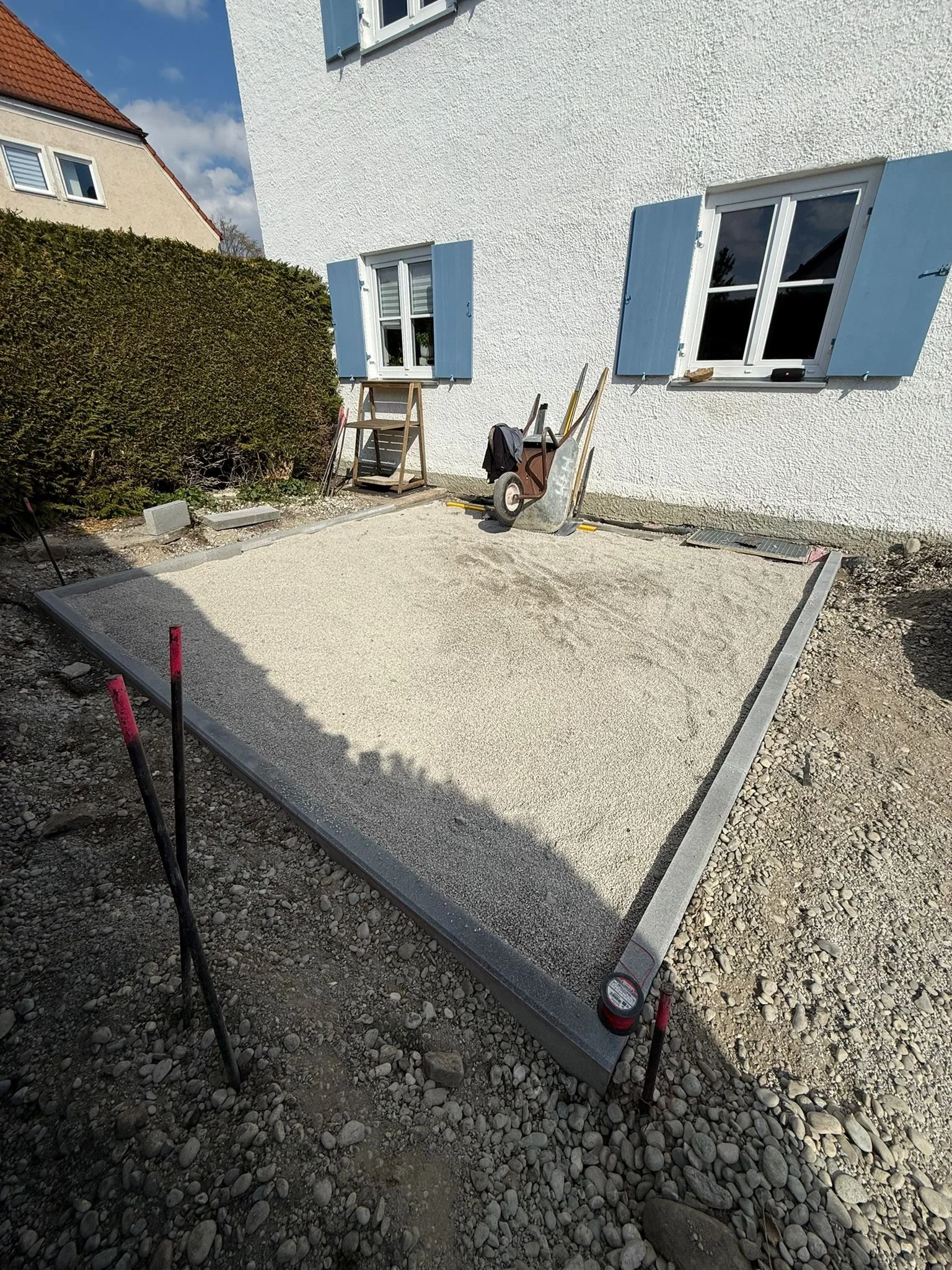 Kies- oder Sandgrund für eine Terrasse vor einem weißen Haus mit blauen Fensterläden, im Bau befindlich.