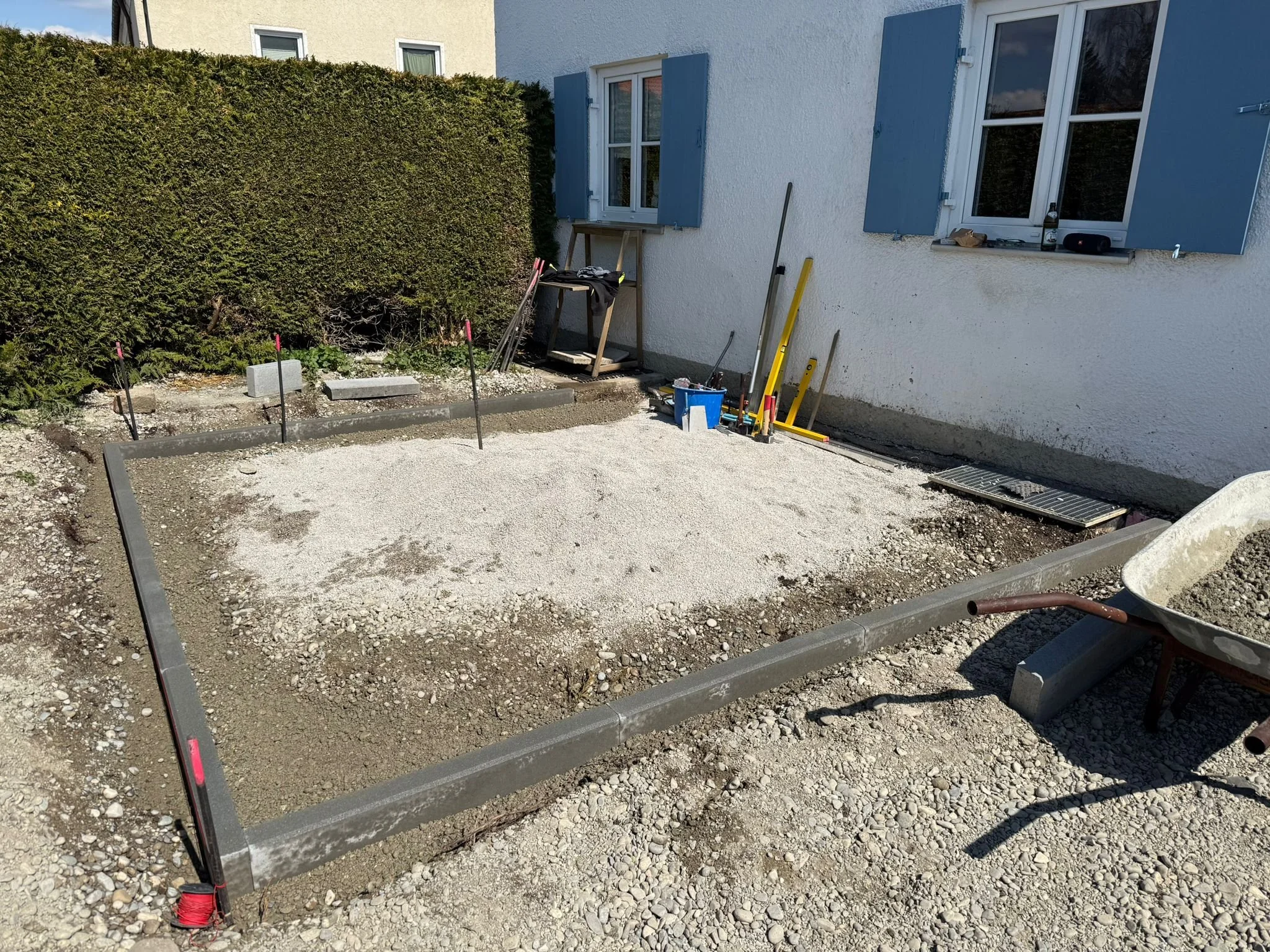 Eine Baustelle im Garten mit vorbereiteter Fläche für eine Terrasse, umgeben von Werkzeugen und einem weißen Haus mit blauen Fensterläden.