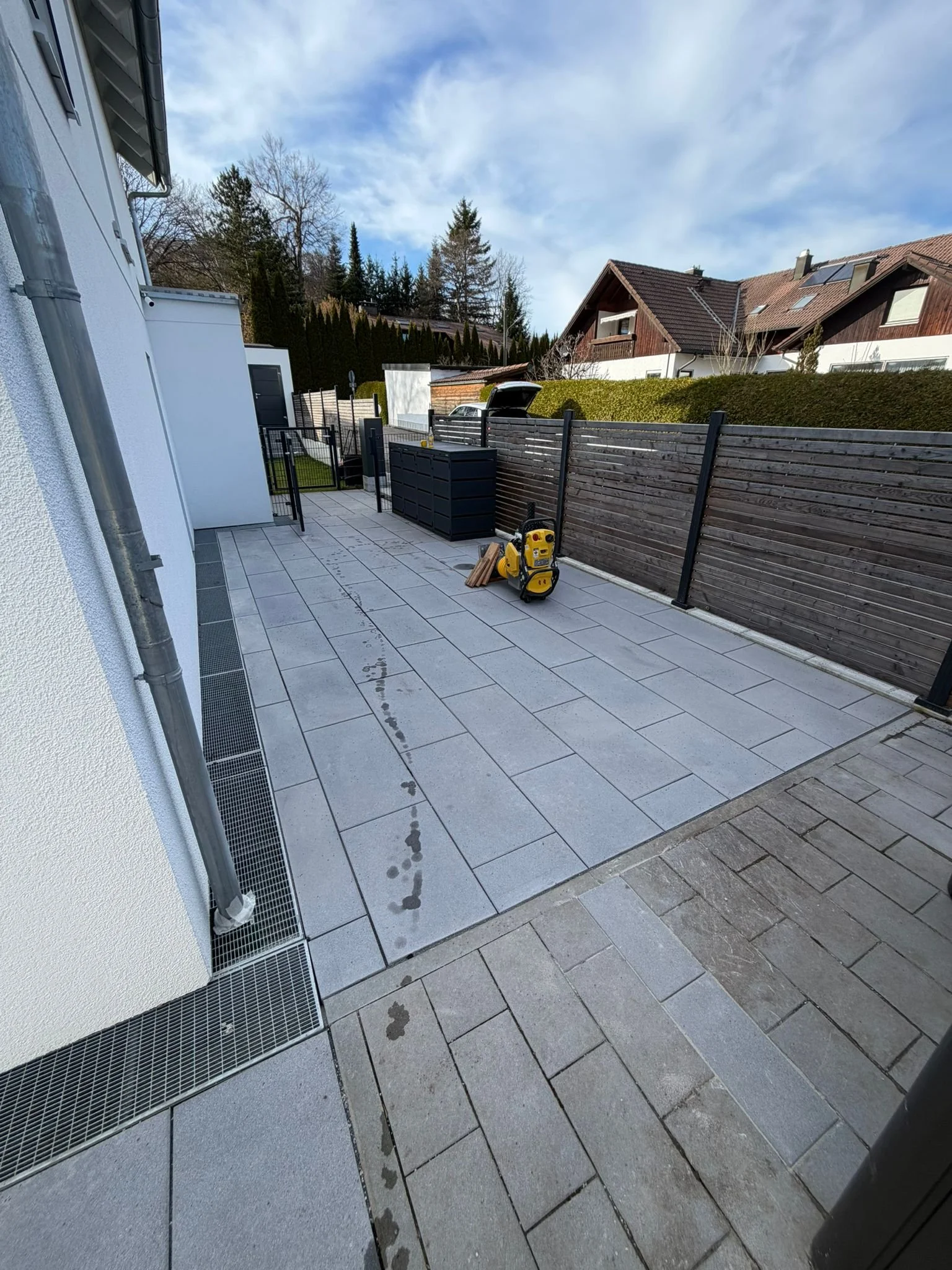 Ein aufgefüllter und geöffneter Müllcontainer steht auf einer Terrasse vor einem hohen Holzzaun. Es gibt ein kleines Holzgestell, eine gelbe elektrische Druckpistole, und einen Stapel Holzstücke, alles auf einem gepflasterten Boden.