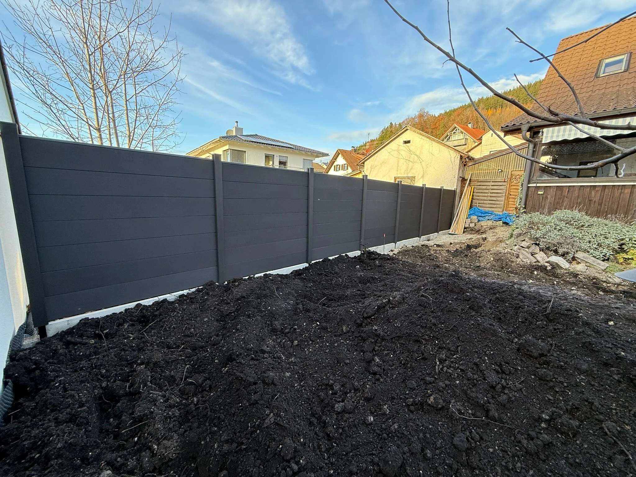 Garten mit dunklem Sichtschutzzaun und ungehobeltem Boden, im Hintergrund sind Häuser mit roten und grauen Dächern und blauer Himmel.