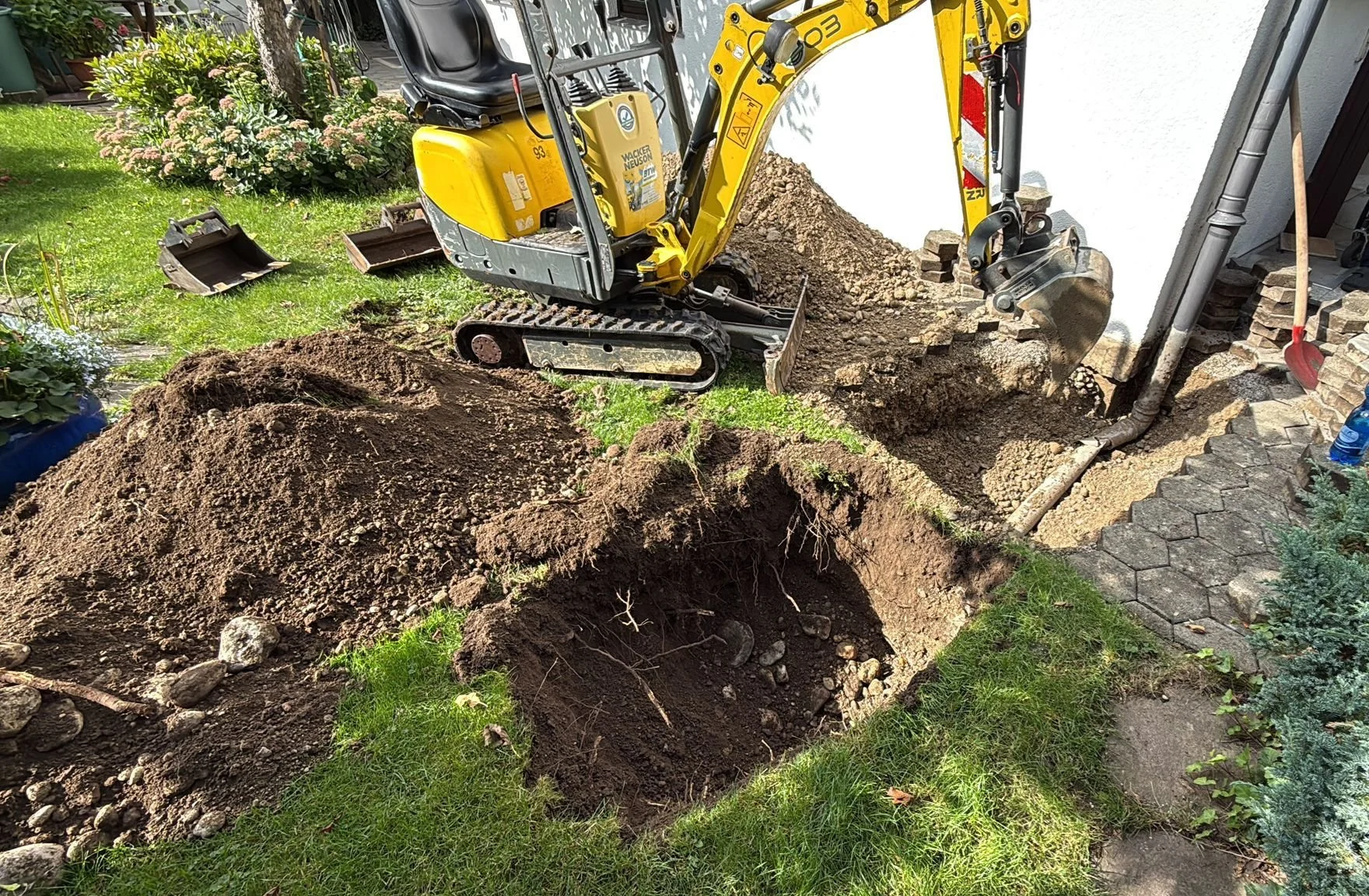 Ein kleiner Bagger gräbt eine Vertiefung im Garten neben einem Haus, mit Erde und Steinen im Arbeitsbereich, umgeben von Rasen und Pflanzen.