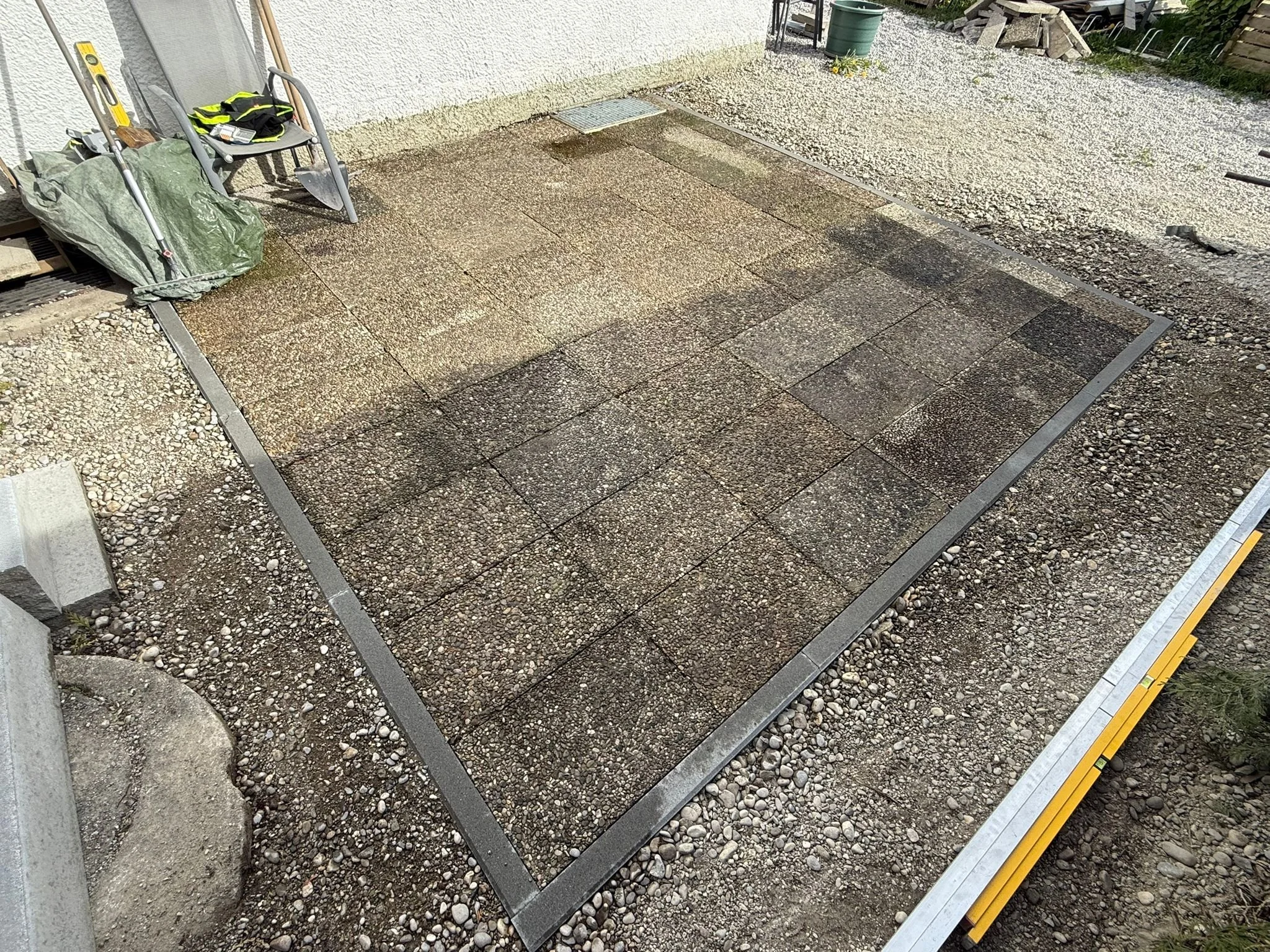 Neue bemusterte Pflastersteine auf dem Boden einer Baustelle, umrissen durch eine Metallbegrenzung, mit Baustoffen im Hintergrund.