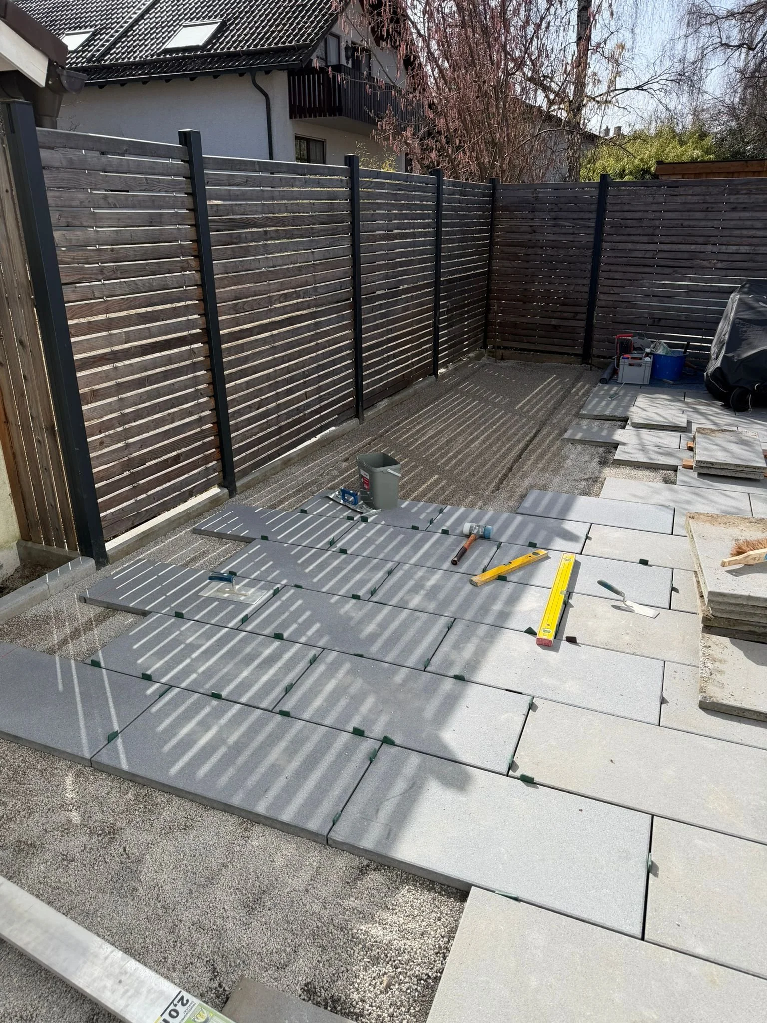 Der Garten wird mit neuen großformatigen terracottafarbenen Natursteinplatten gepflastert, während die alten Pflastersteine herausgenommen wurden. Ein Zaun aus dunklem Holz gibt die Begrenzung des Gartens ab.