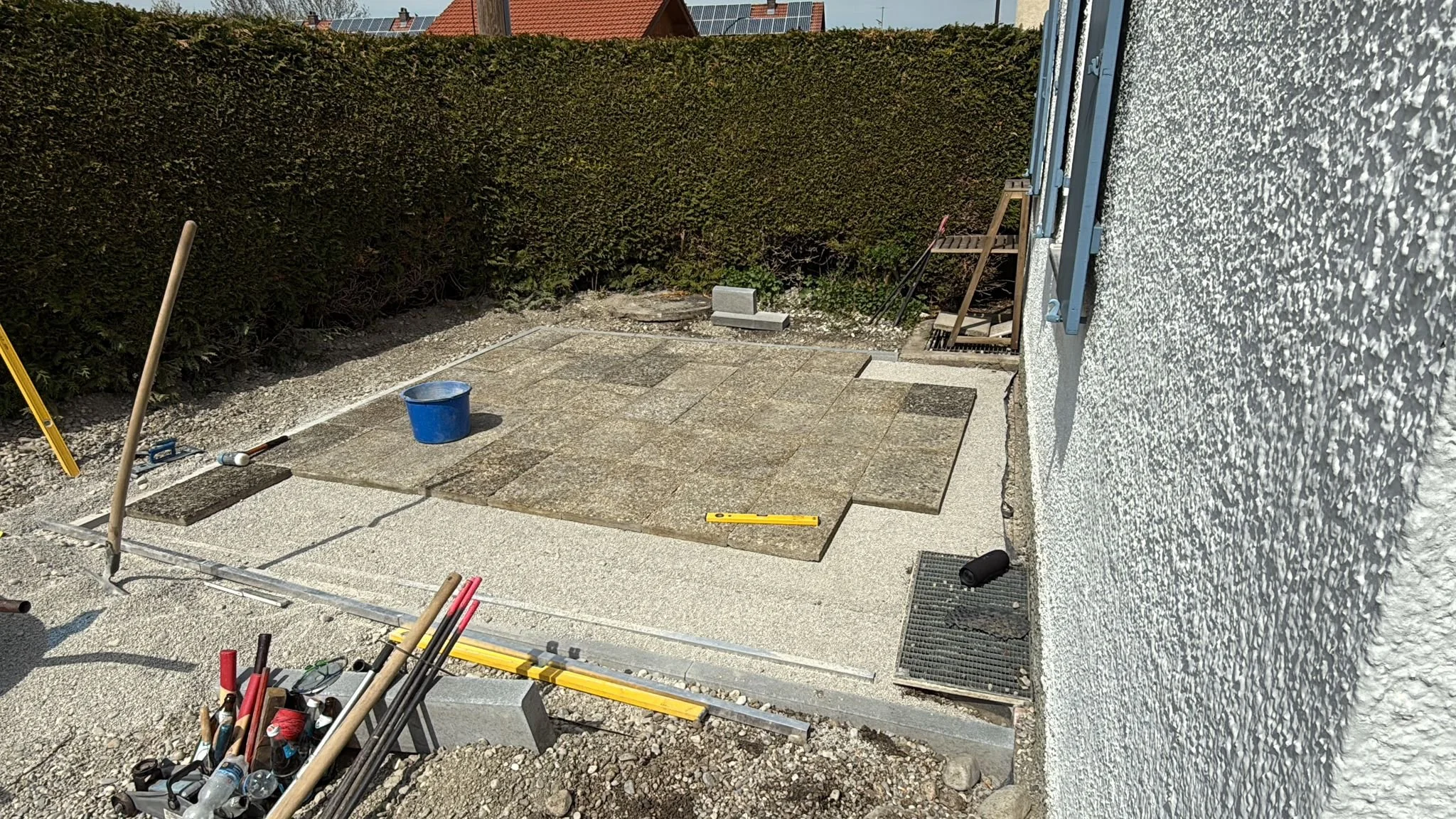 Auf einer Terrasse werden Pflastersteine verlegt. Es gibt Werkzeuge, eine Wasserwaage, eine blaue Eimer, einen Möbelstuhl und einen Rollladenschalter an der Wand.