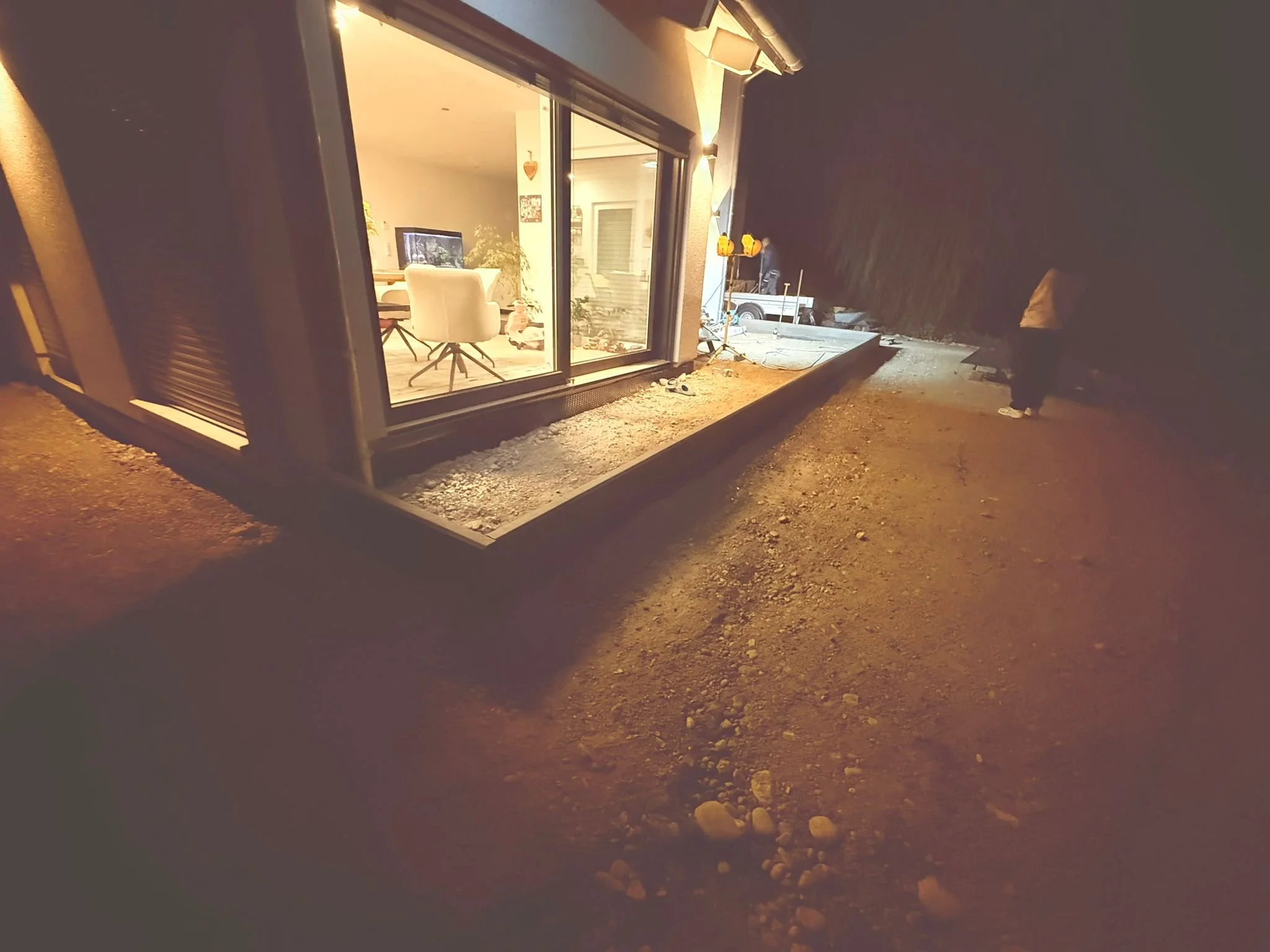 Blick auf ein modernes Haus mit großer Fensterverglasung bei Nacht, im Inneren sind ein Esstisch und mehrere Stühle sichtbar, der Außenbereich ist noch im Bau oder Renovierung, mit Erde und Steinen vor dem Haus