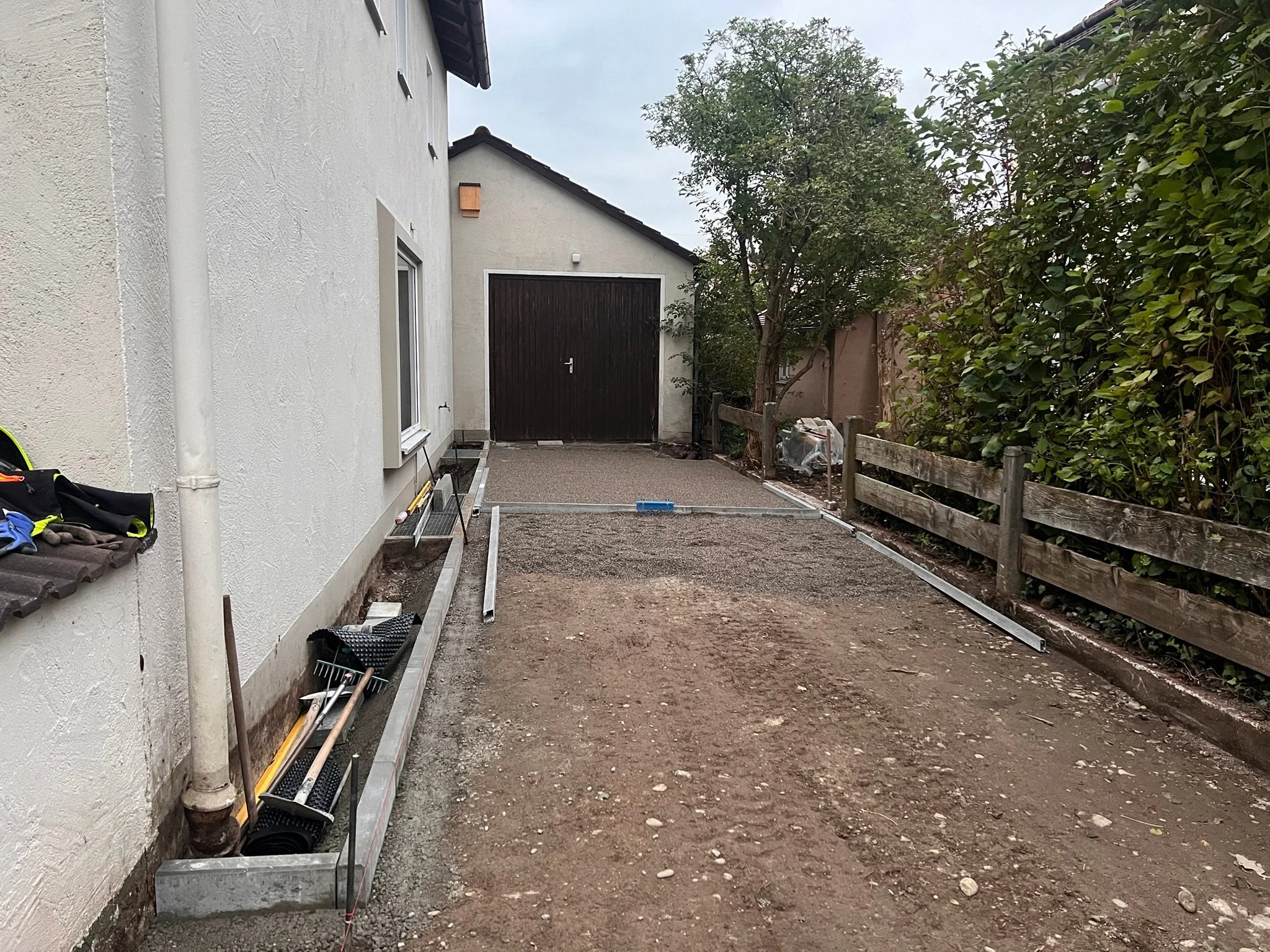 Ein Gartenweg wird neu gestaltet, mit Werkzeugen und Baumaterialien bereitgestellt, an einem Haus mit weißer Wand, im Hintergrund ein Garagentor und Bäume.