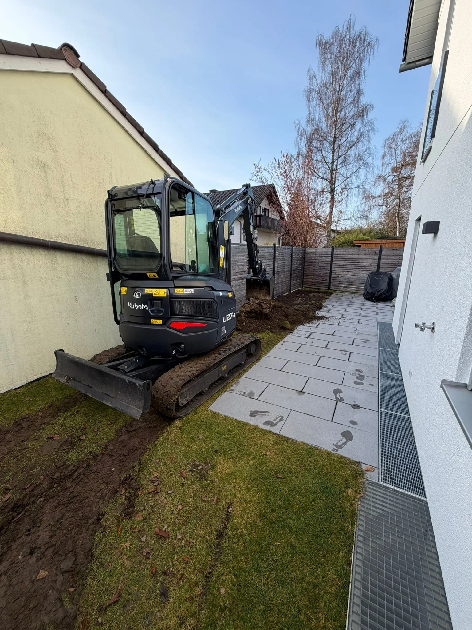 Ein kleines schwarzes Baggergerät arbeitet in einem Garten, um den Boden für eine Pflasterung vorzubereiten, mit einem Zaun, Häusern und Bäumen im Hintergrund, bei sonnigem Himmel.