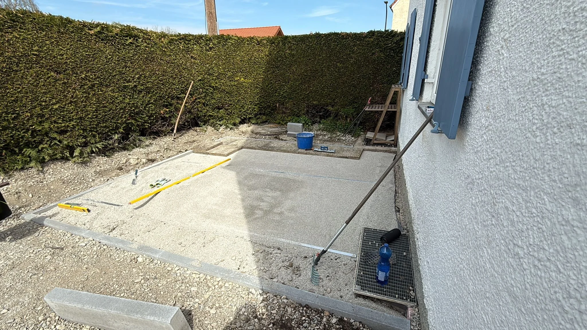 Bauarbeiten im Garten mit Sand, Werkzeugen und Baumaterialien vor einem weißen Haus, um einen neuen Gehweg zu bauen.