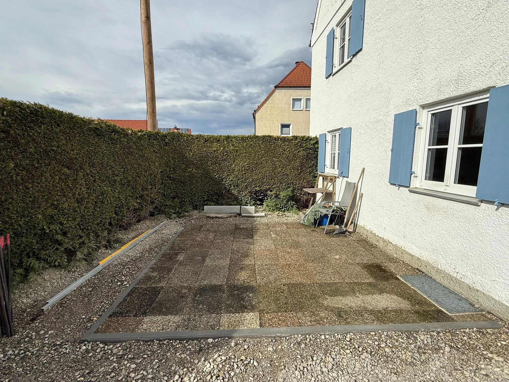Gartenfläche vor einem weißen Haus mit blauen Fensterläden, teilweise eingezäunt, mit Gartenarbeitzeugen und Baumaterialien, bei bewölktem Himmel.